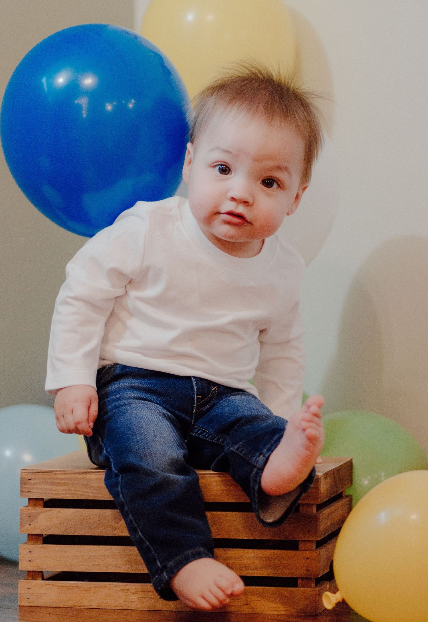 Adorable baby with Down syndrome sitting on wooden crate surrounded by colorful birthday balloons.