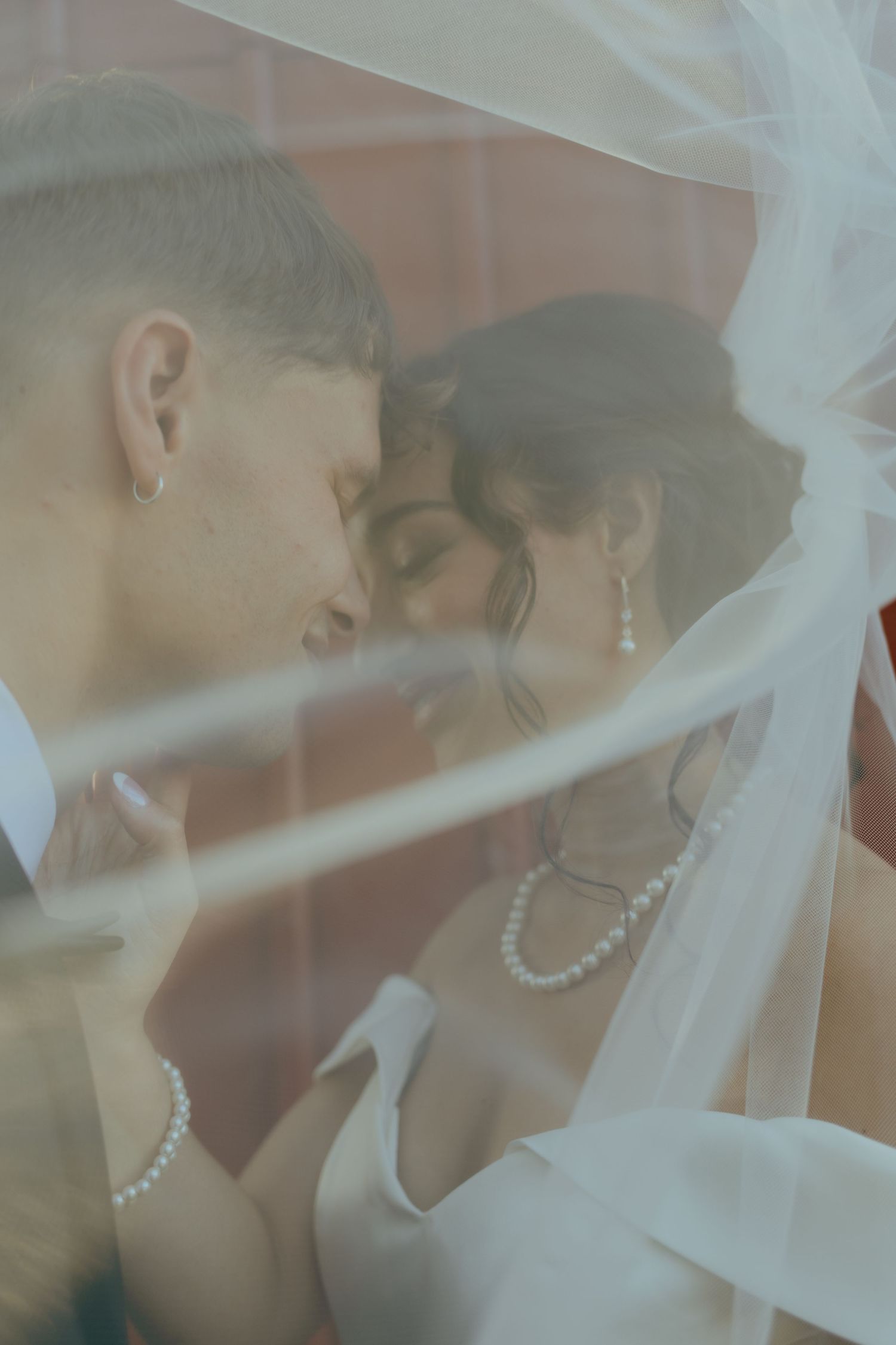 Intimate bride and groom almost kissing beneath a sheer wedding veil, wearing pearl jewelry and wedding attire.