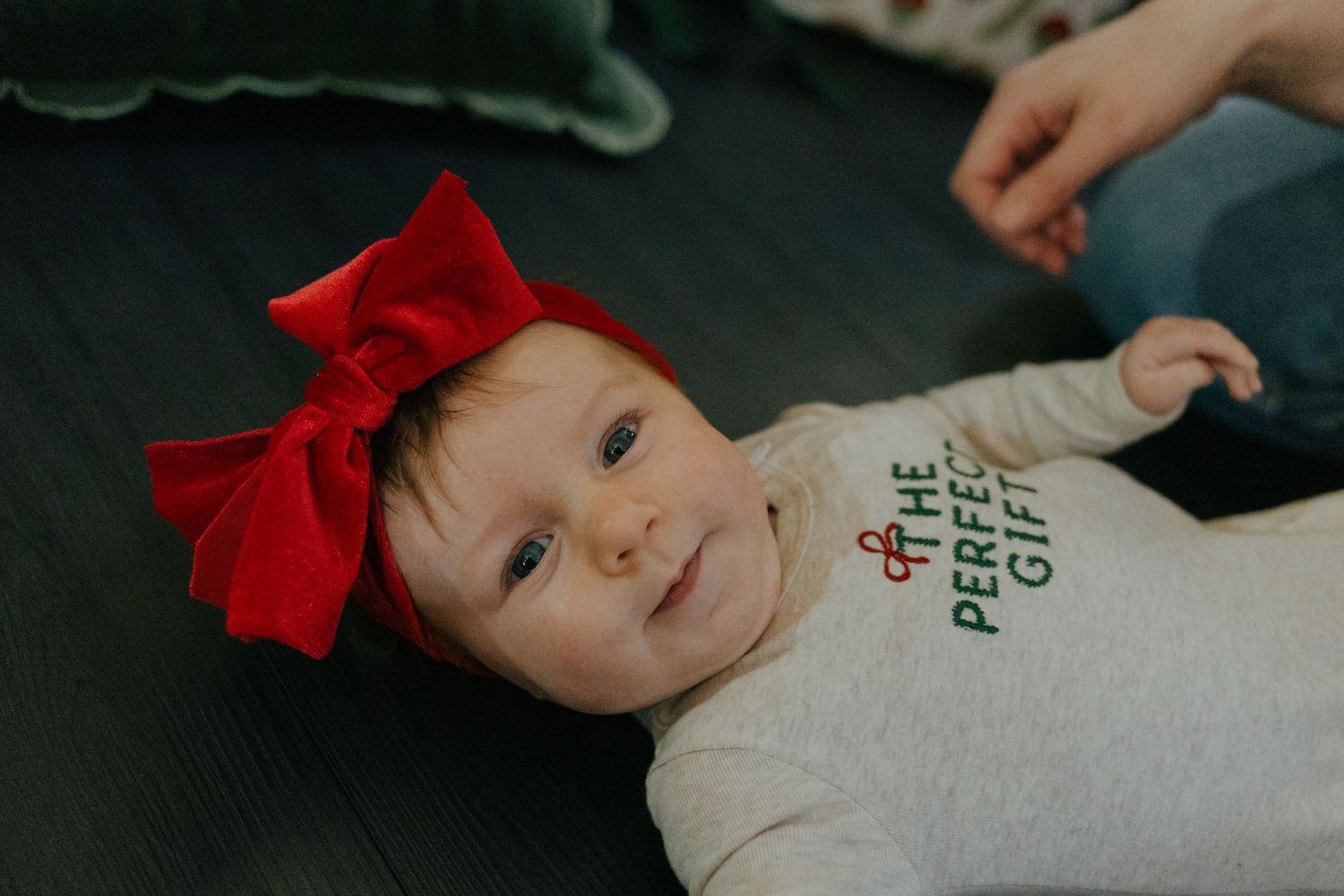 Adorable baby wearing red bow headband and The Perfect Gift onesie lying on dark floor.