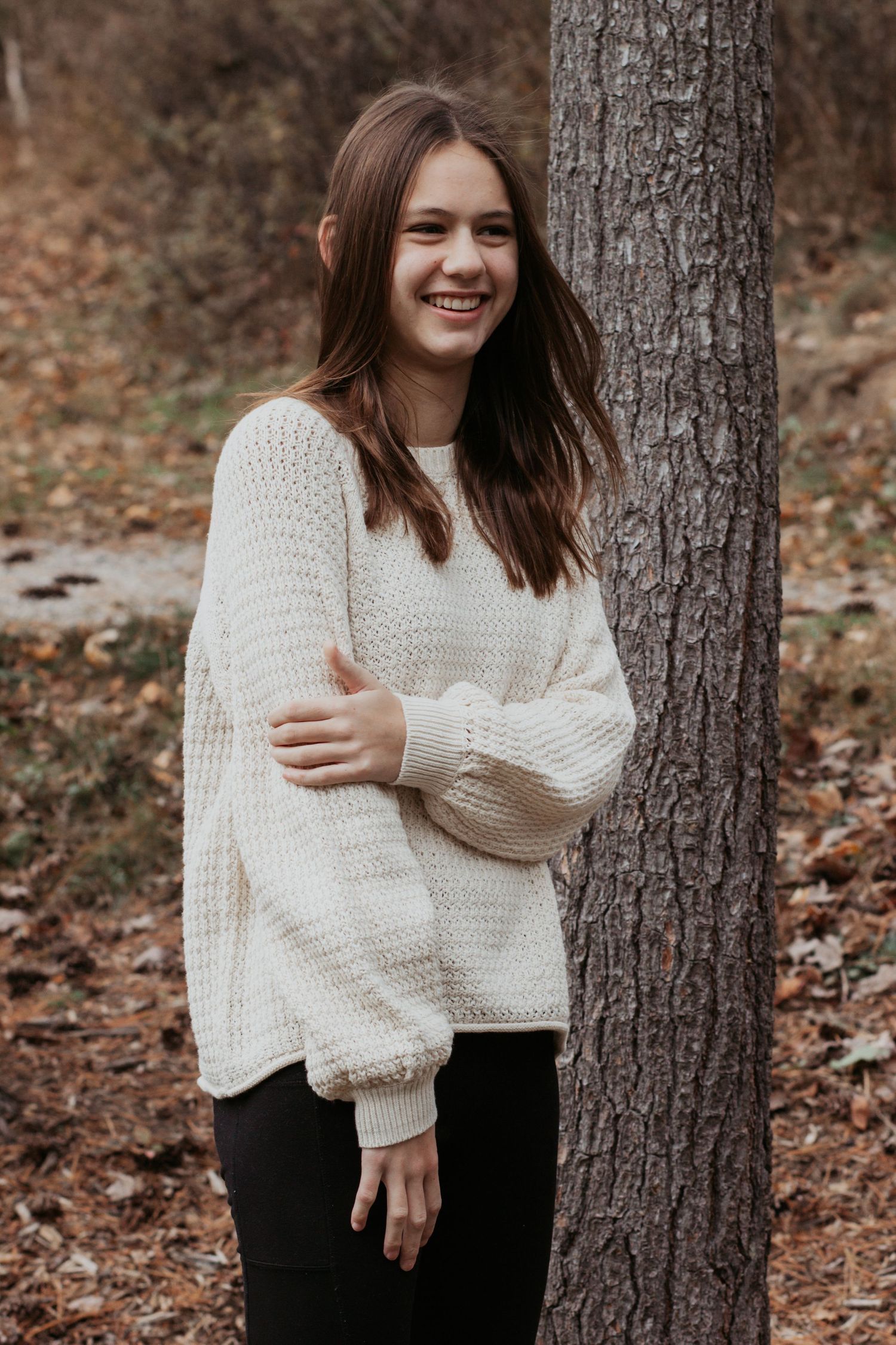 Smiling teenage girl in cream knit sweater leaning against tree in fall forest.