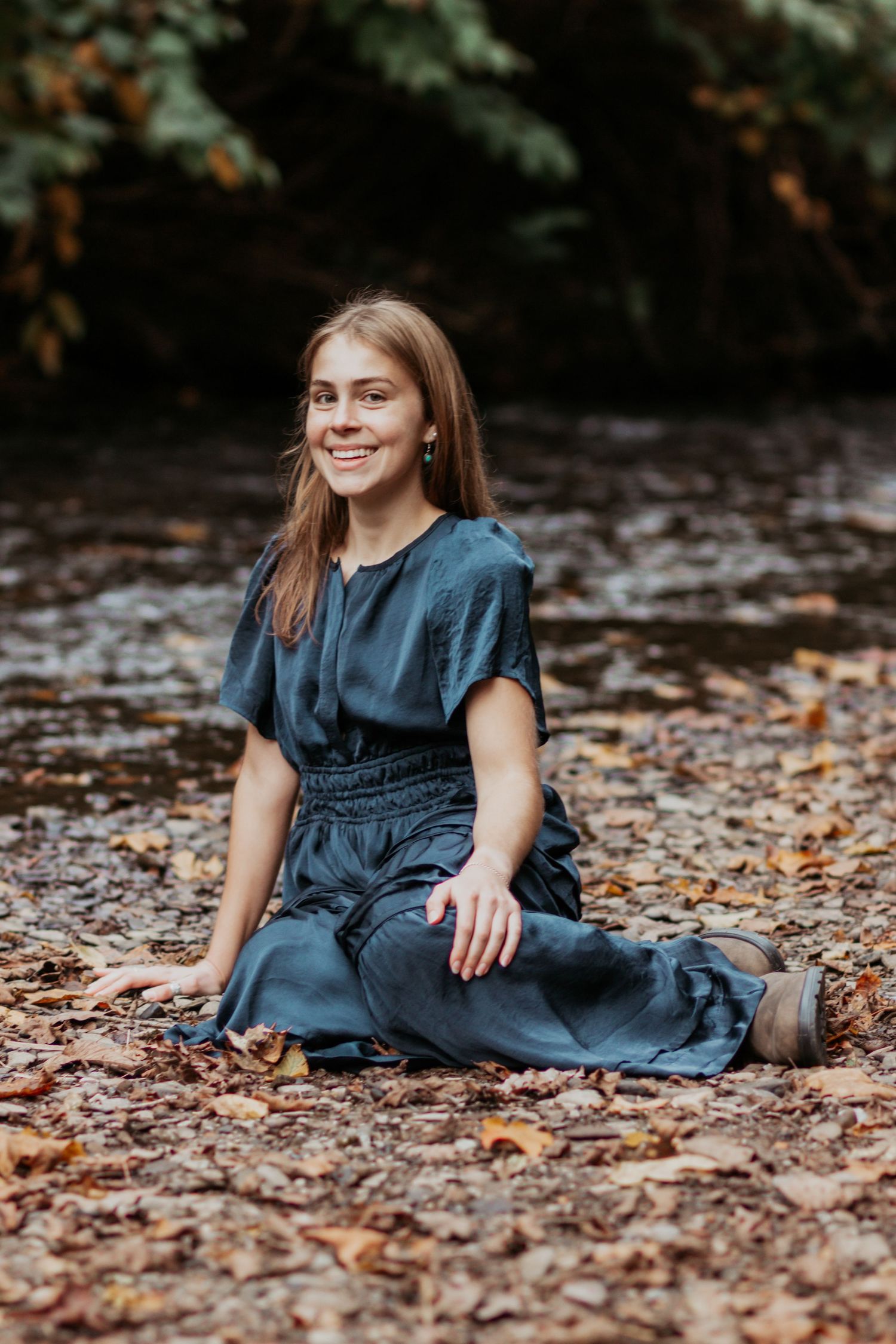 Smiling young woman in teal dress sitting on autumn leaves near a creek.