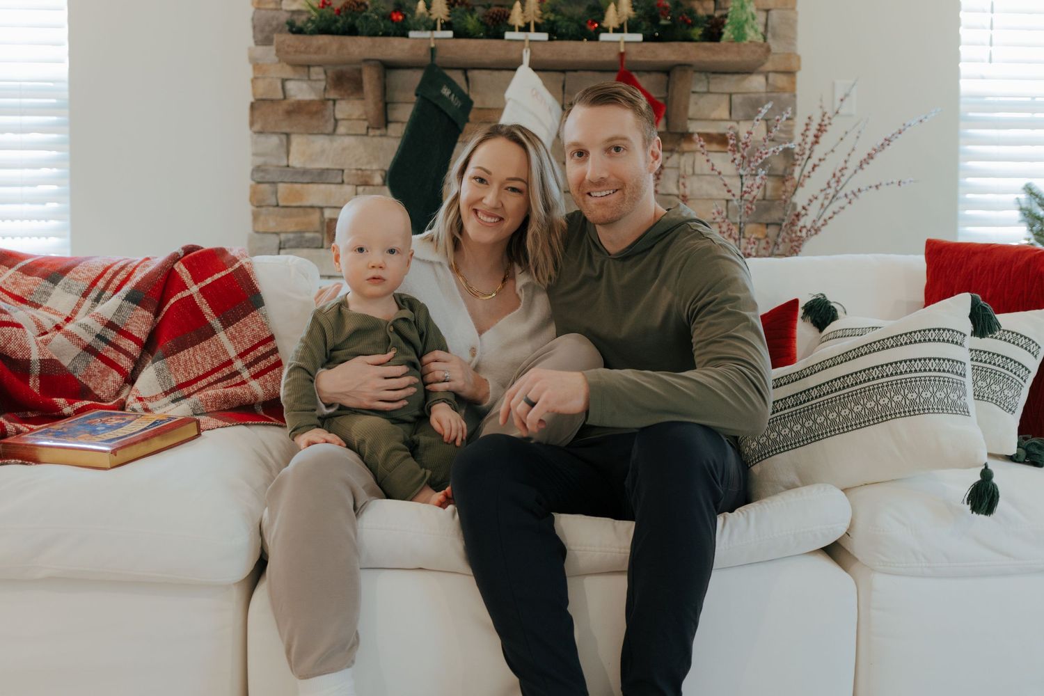 Smiling couple with baby in holiday outfits sit together on white couch with Christmas decorations.
