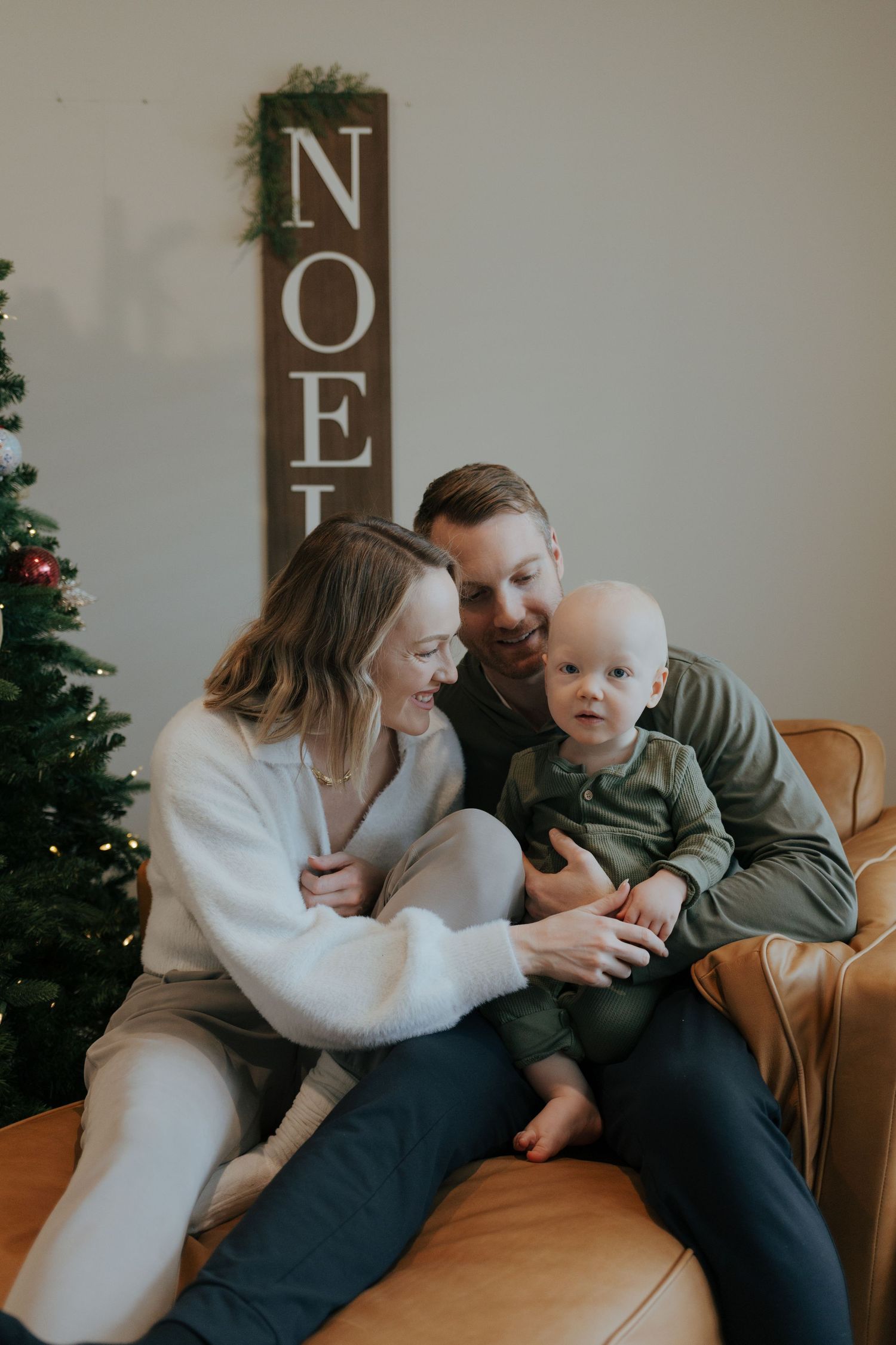 Young family with baby boy relaxes on leather couch next to Christmas tree and NOEL sign.