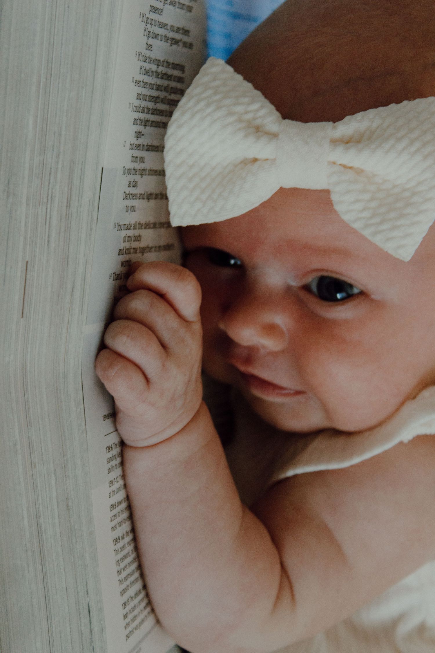 Newborn baby girl with cream bow headband lying next to an open book, grasping pages with tiny hand.