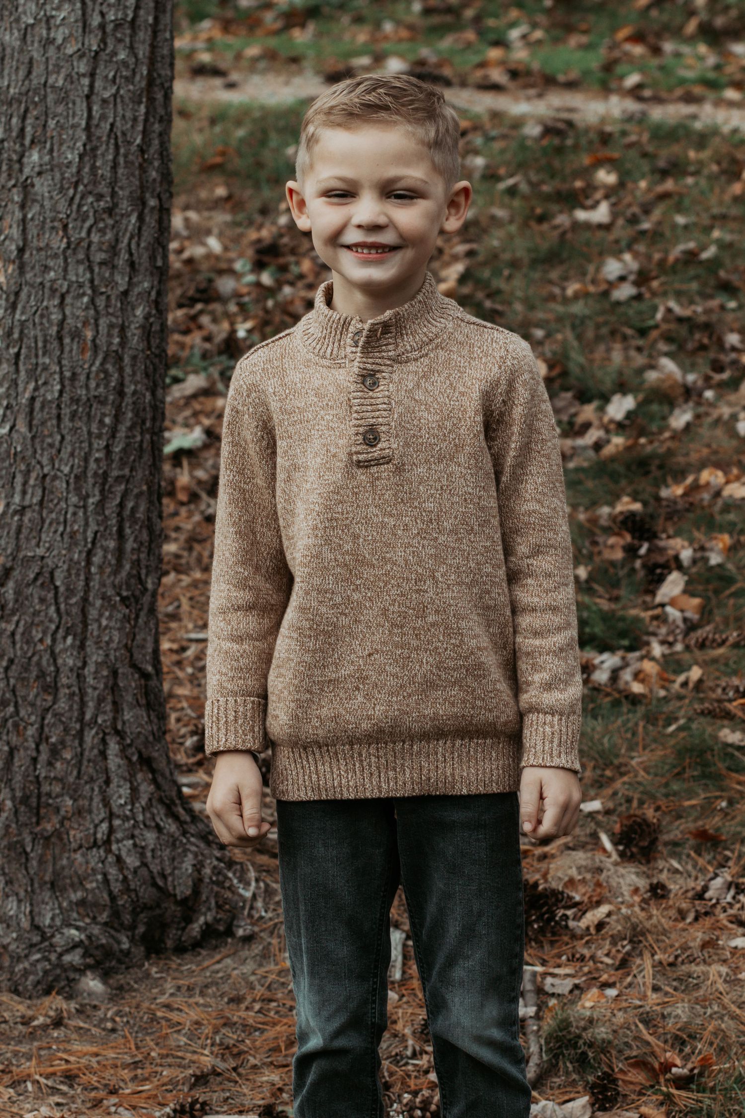 Young boy in tan button-neck sweater smiling beside tree in fall forest.