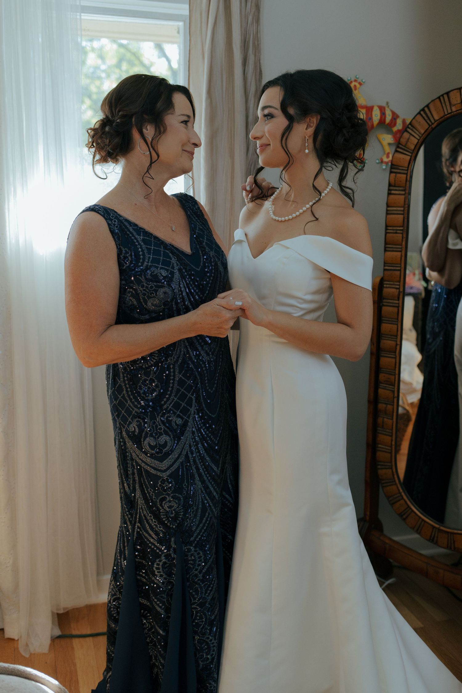Bride in off-shoulder white gown sharing a tender moment with woman in navy beaded dress.