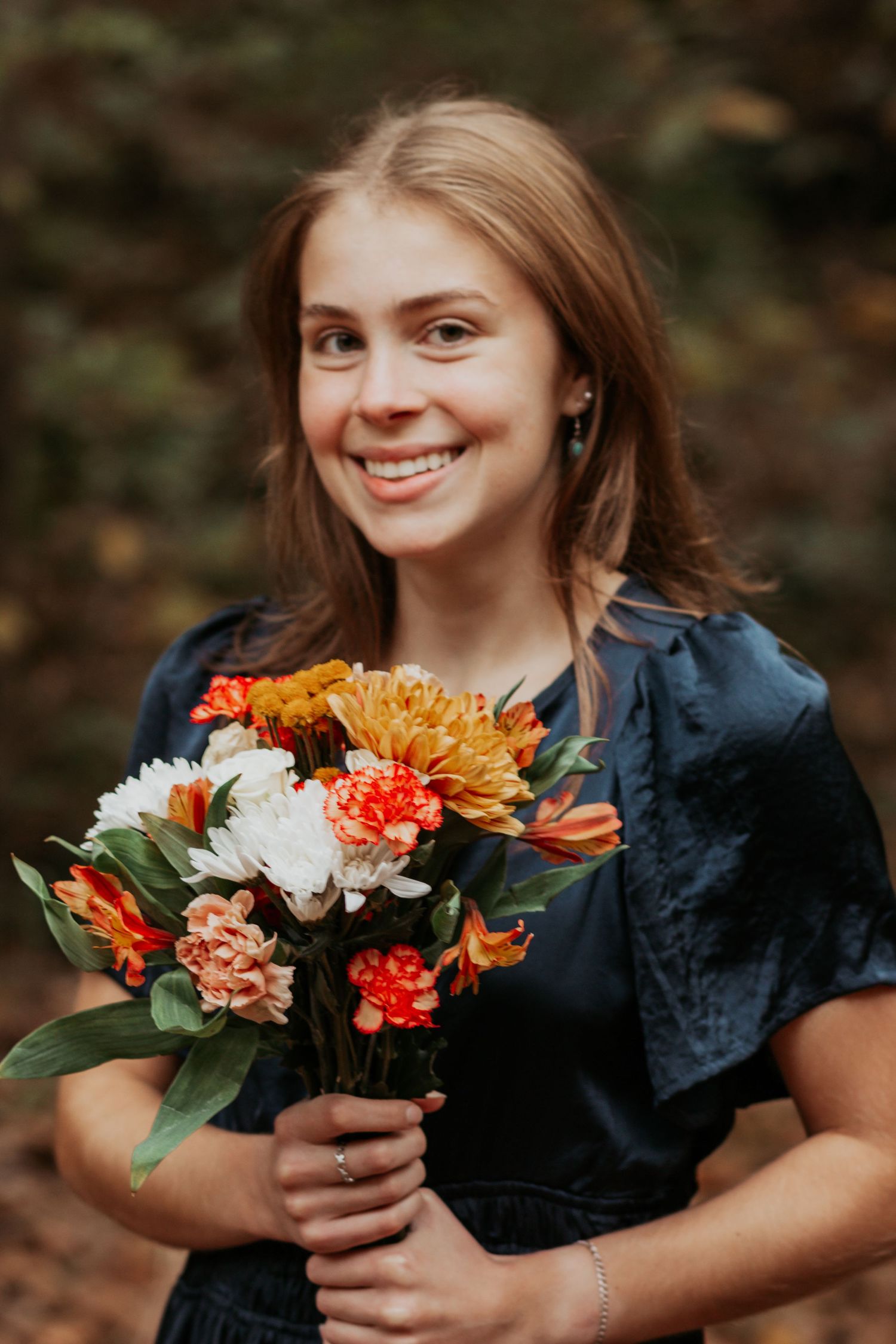 Smiling young woman in dark dress holds colorful autumn bouquet of orange and white flowers outdoors.