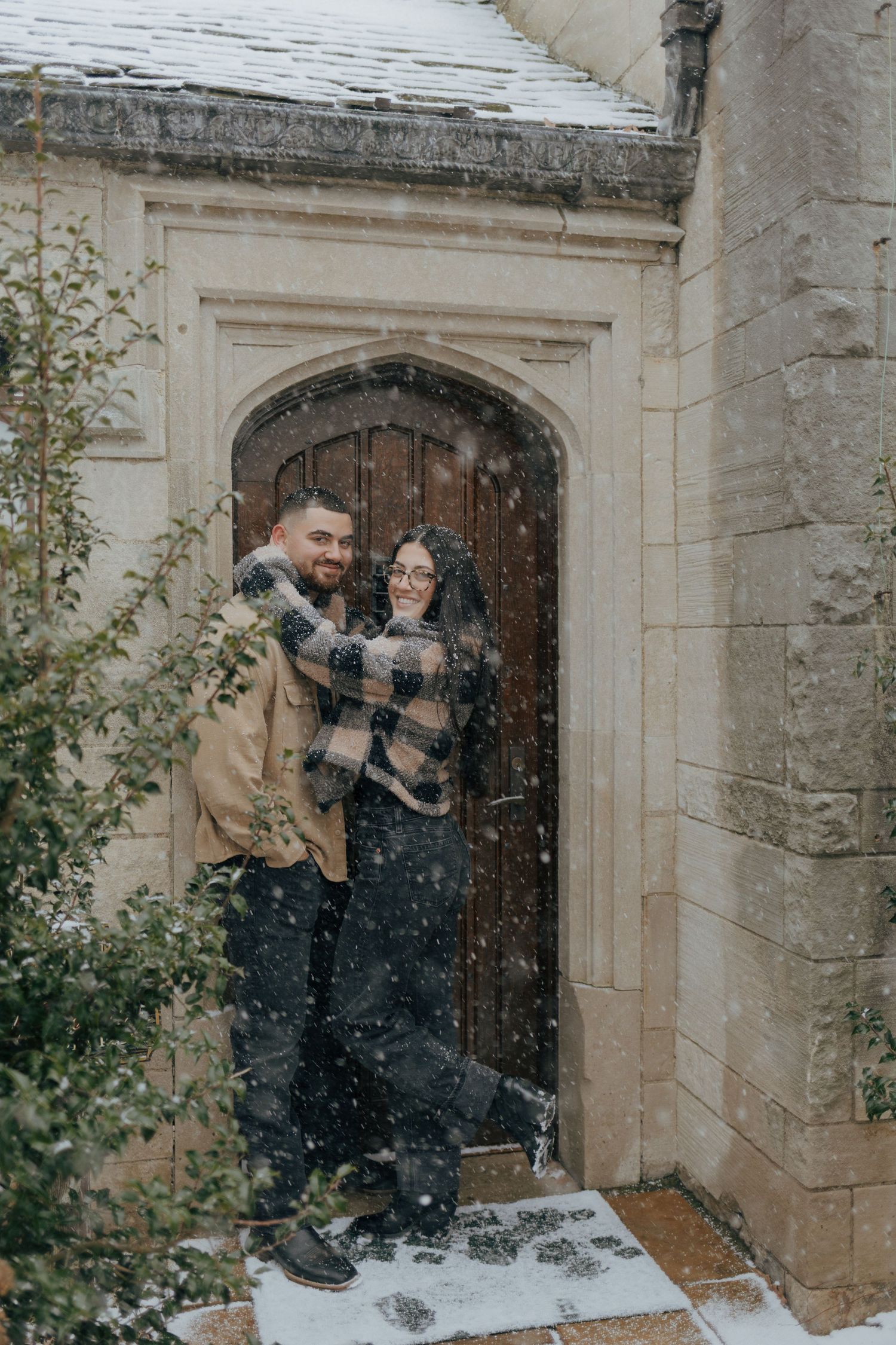 Couple laughing together in snowfall at an arched stone doorway of a historic building in Pittsburgh.