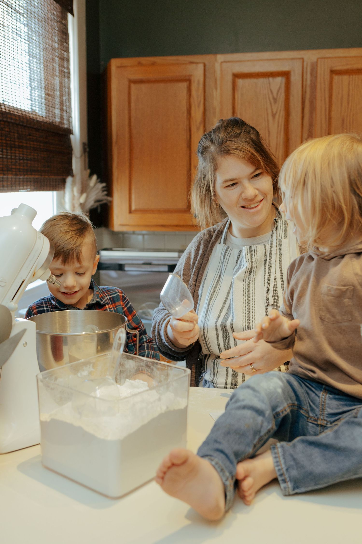 Happy mother smiling while baking with two young children sitting on kitchen counter with flour.