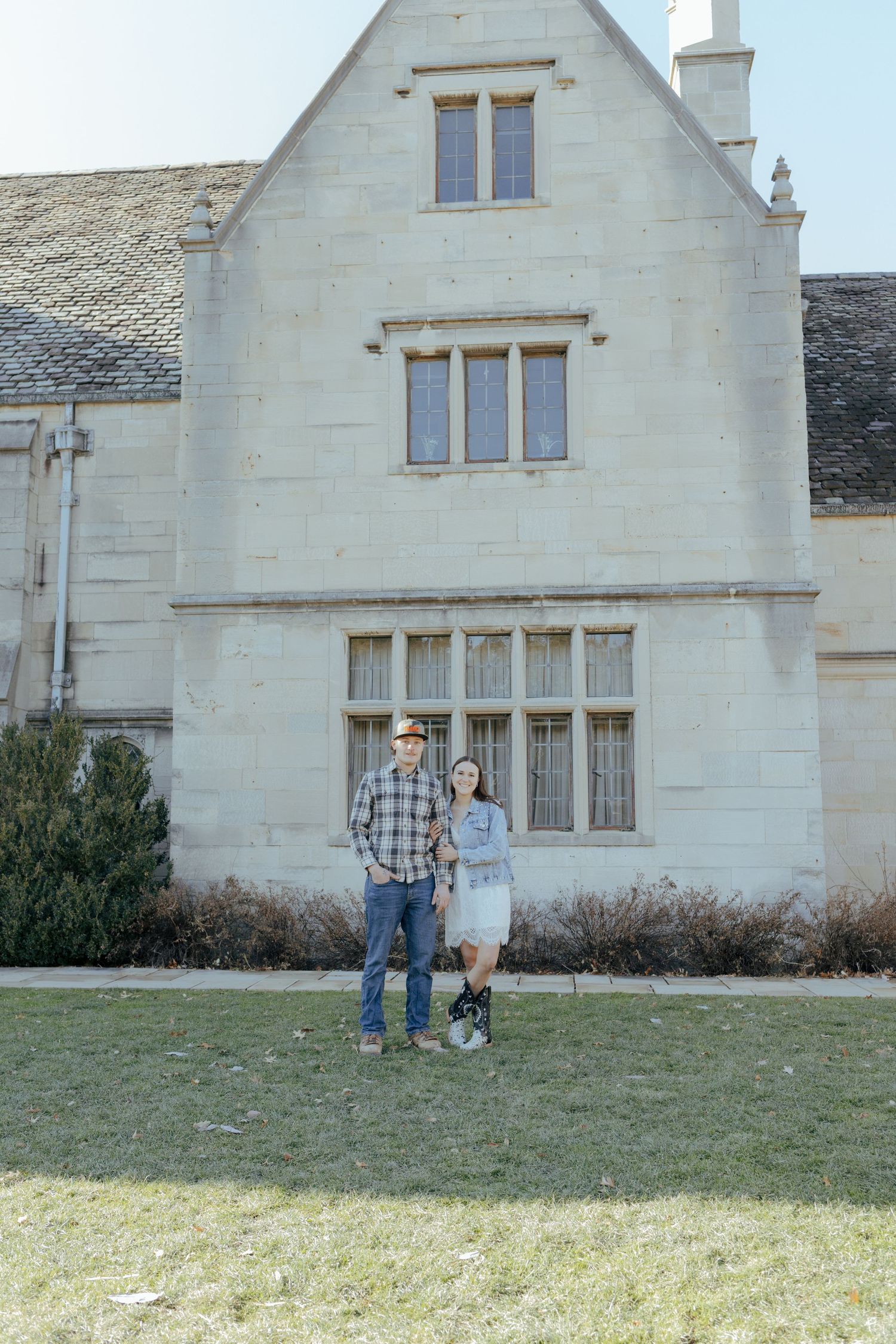 Couple standing together on green lawn in front of a large historic stone manor building.