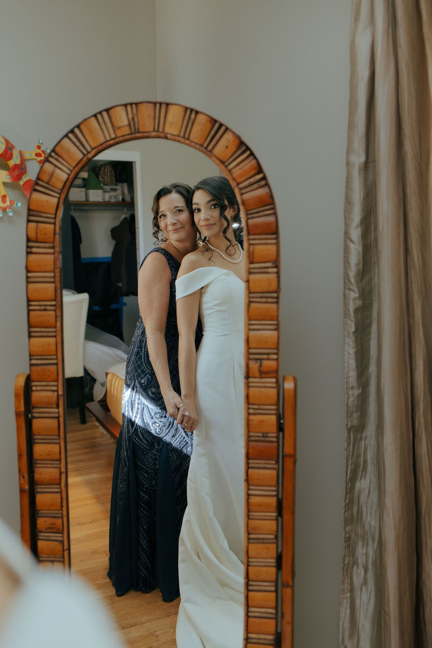 Bride in white off-shoulder gown poses with woman in black dress before arched mirror.