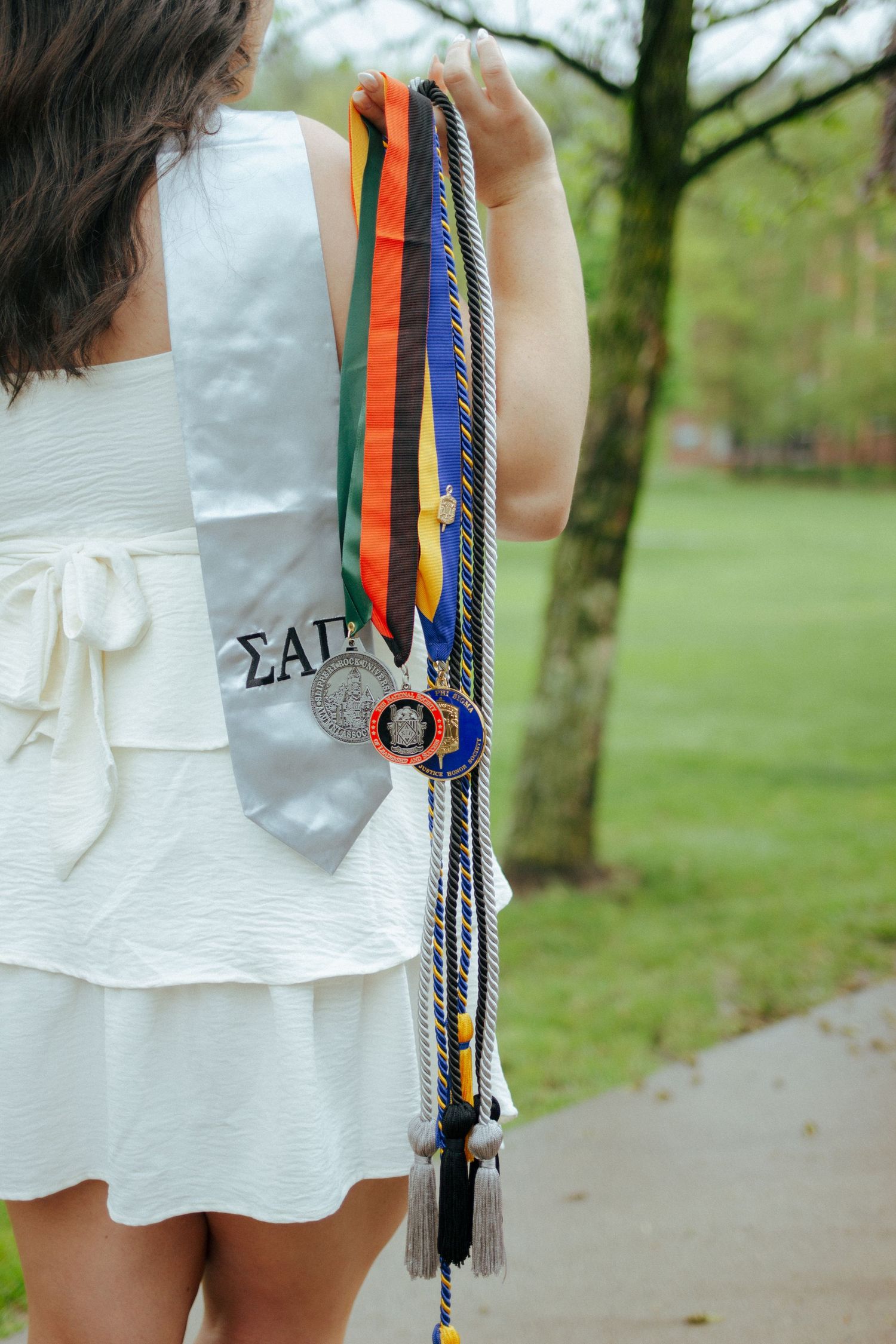 Graduate displays colorful honor cords and medals on a white sorority sash outdoors.