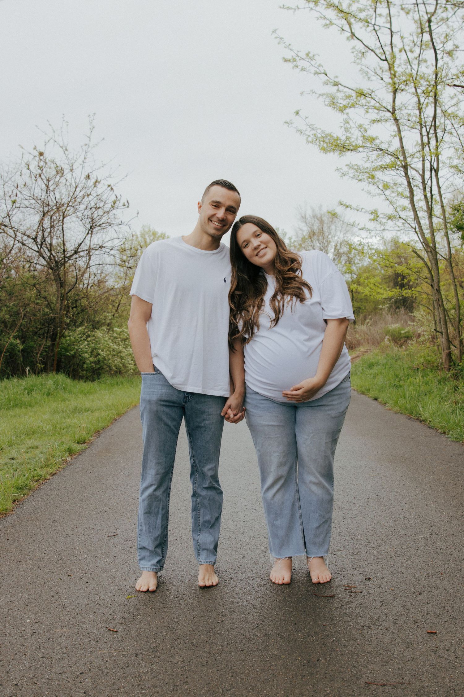 Happy expecting couple in matching white shirts holding hands on a path at Settlers Cabin Park