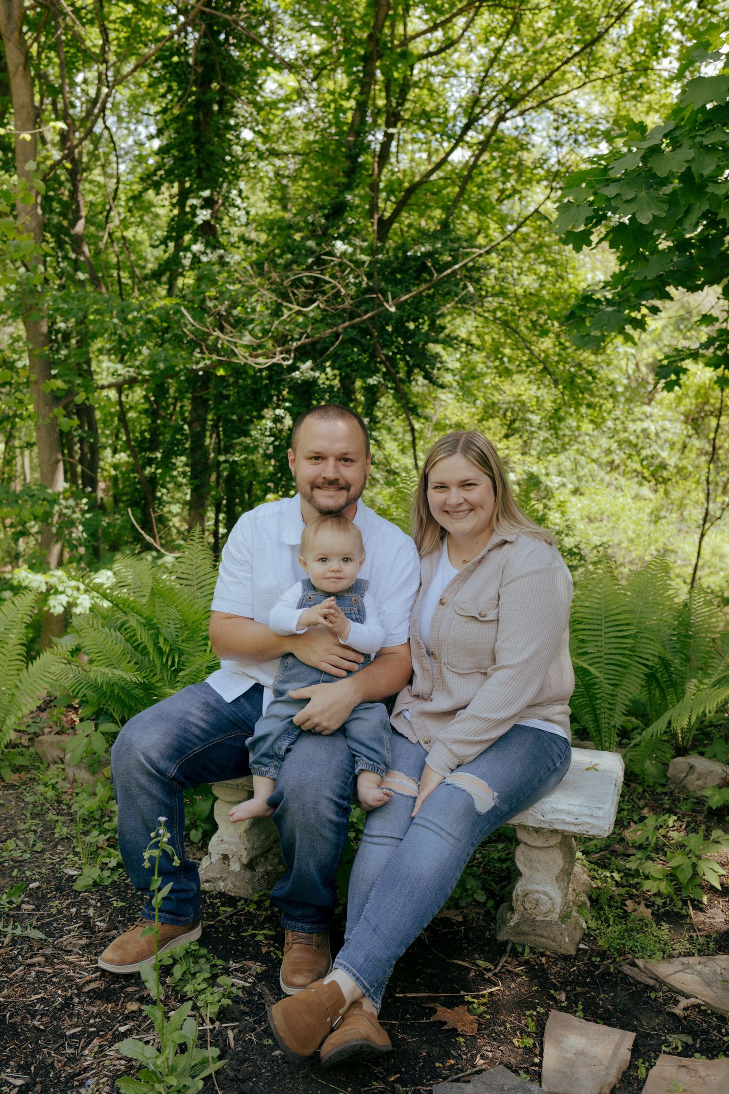 Smiling family of three sitting on a stone bench surrounded by lush green ferns in a garden.