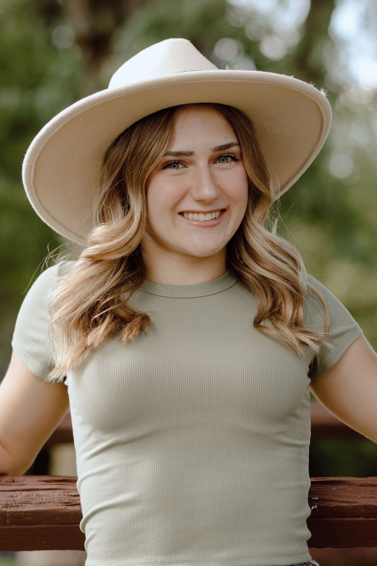 Smiling blonde teen in sage ribbed tee and cream wide-brim hat at wooden fence.