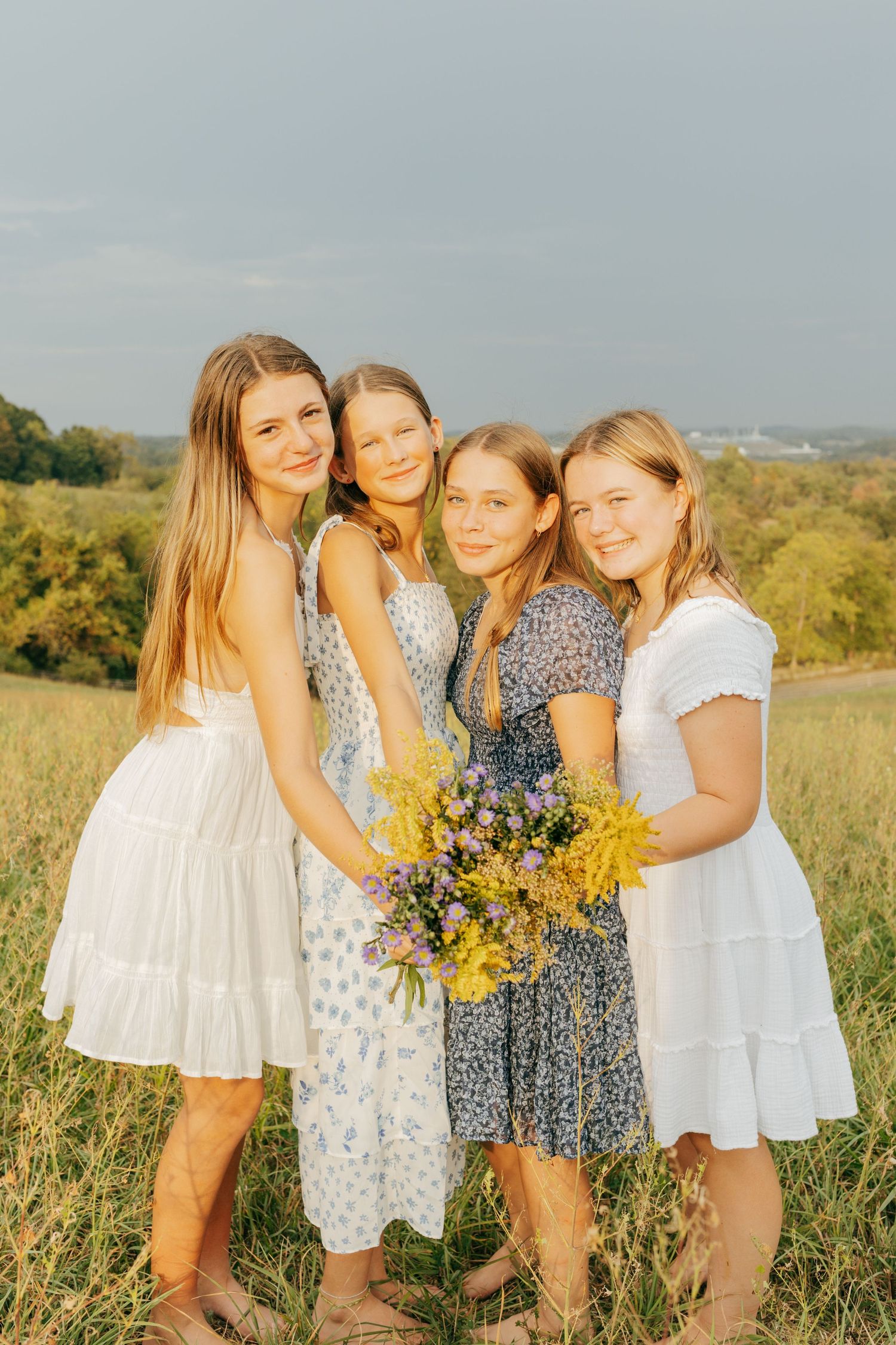 Four girls in summer dresses hold wildflowers in a sunlit meadow.
