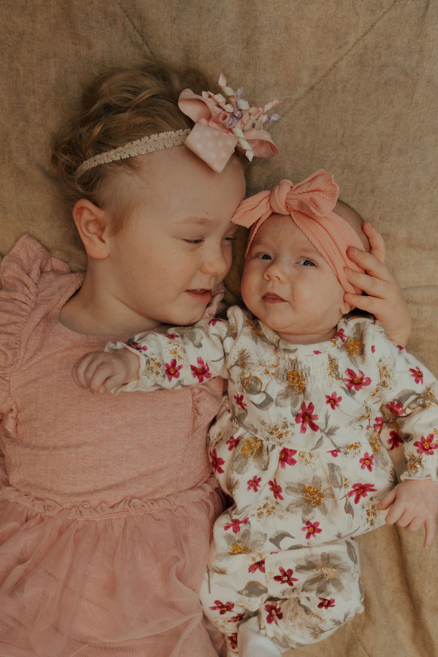 Two young sisters in pink outfits lay together, older toddler gently embracing newborn baby sister.