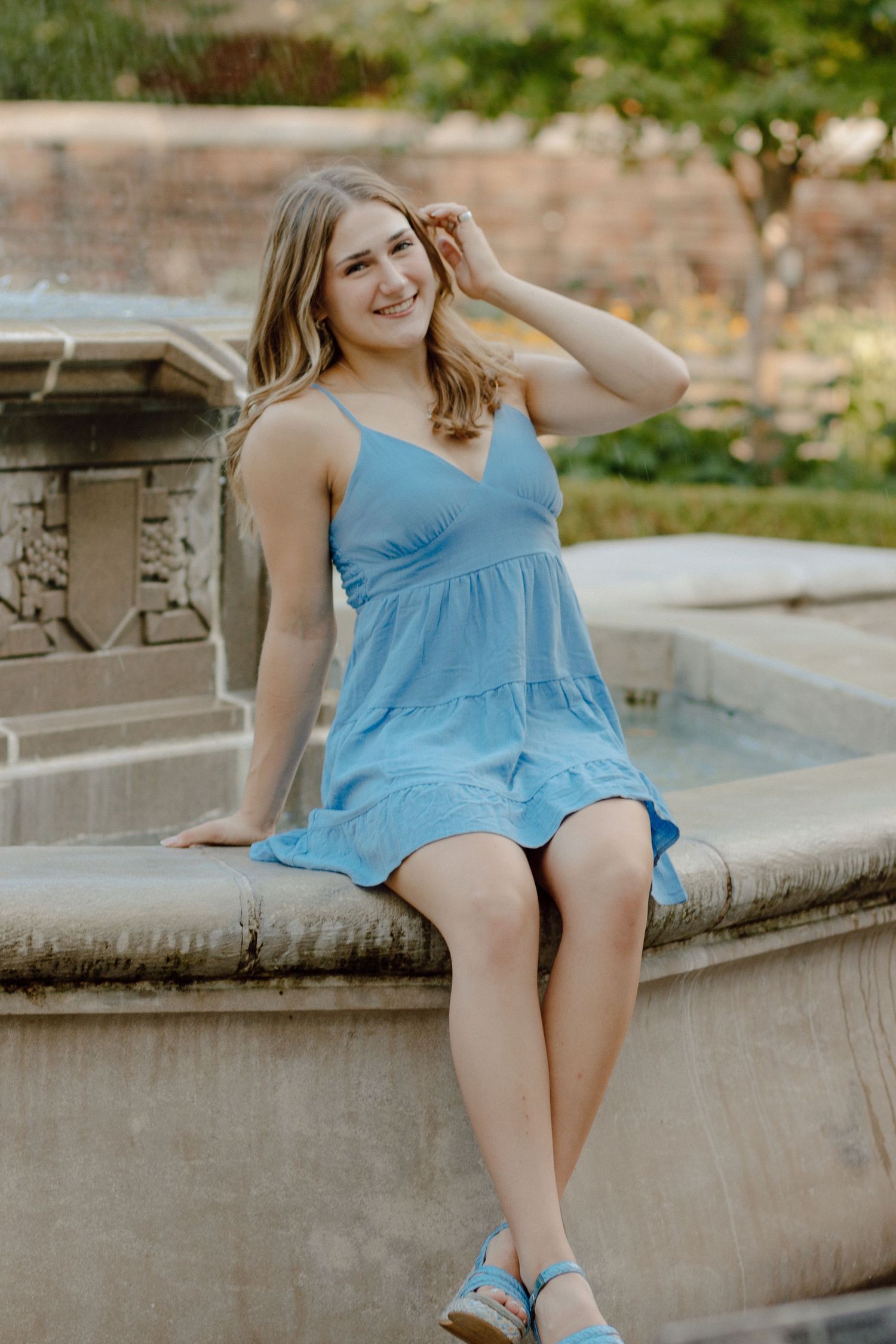 Young woman in light blue sundress sits on stone fountain edge smiling in outdoor garden setting.