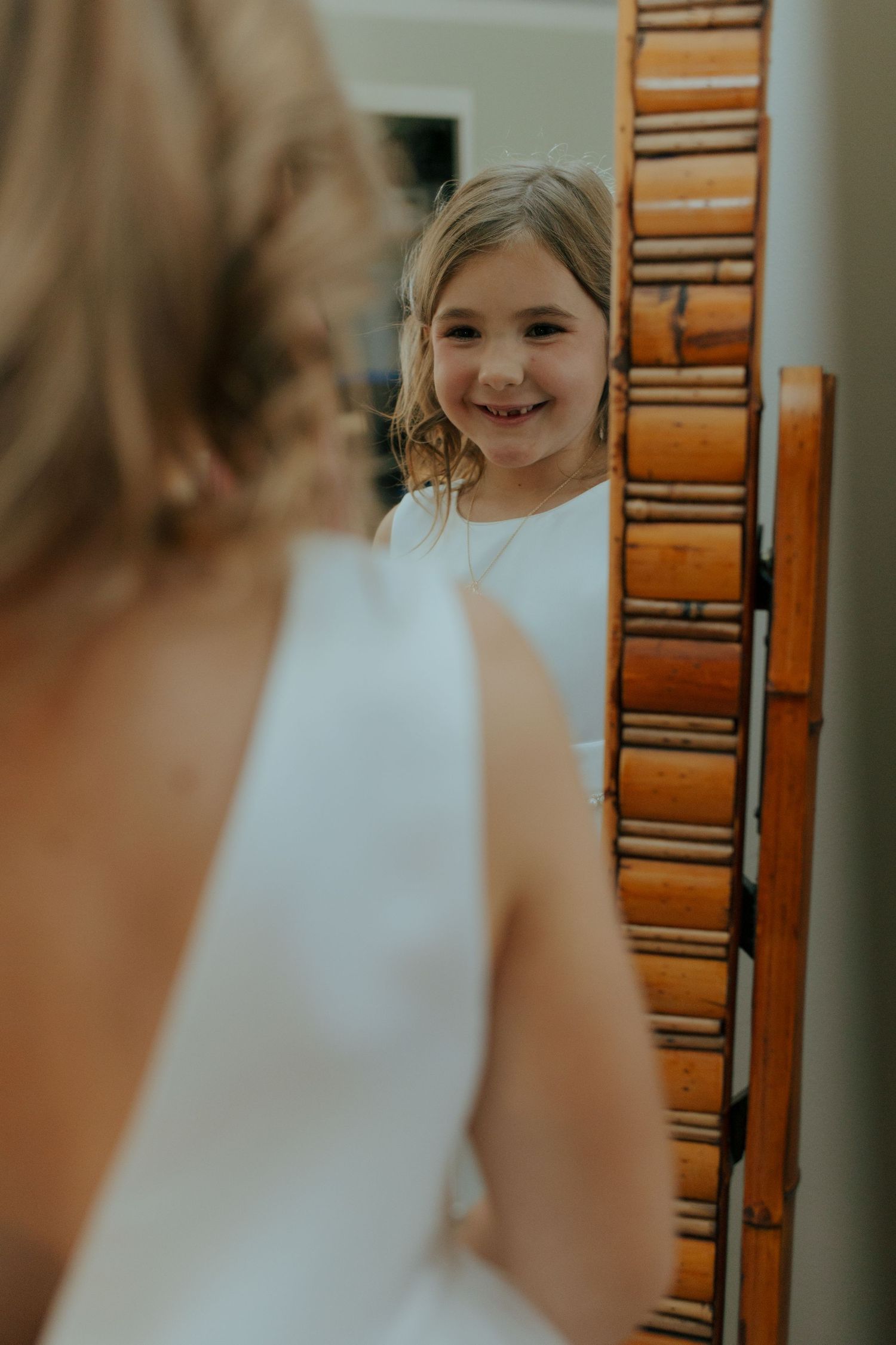 Smiling flower girl in white dress reflected in a bamboo-framed mirror beside the bride.
