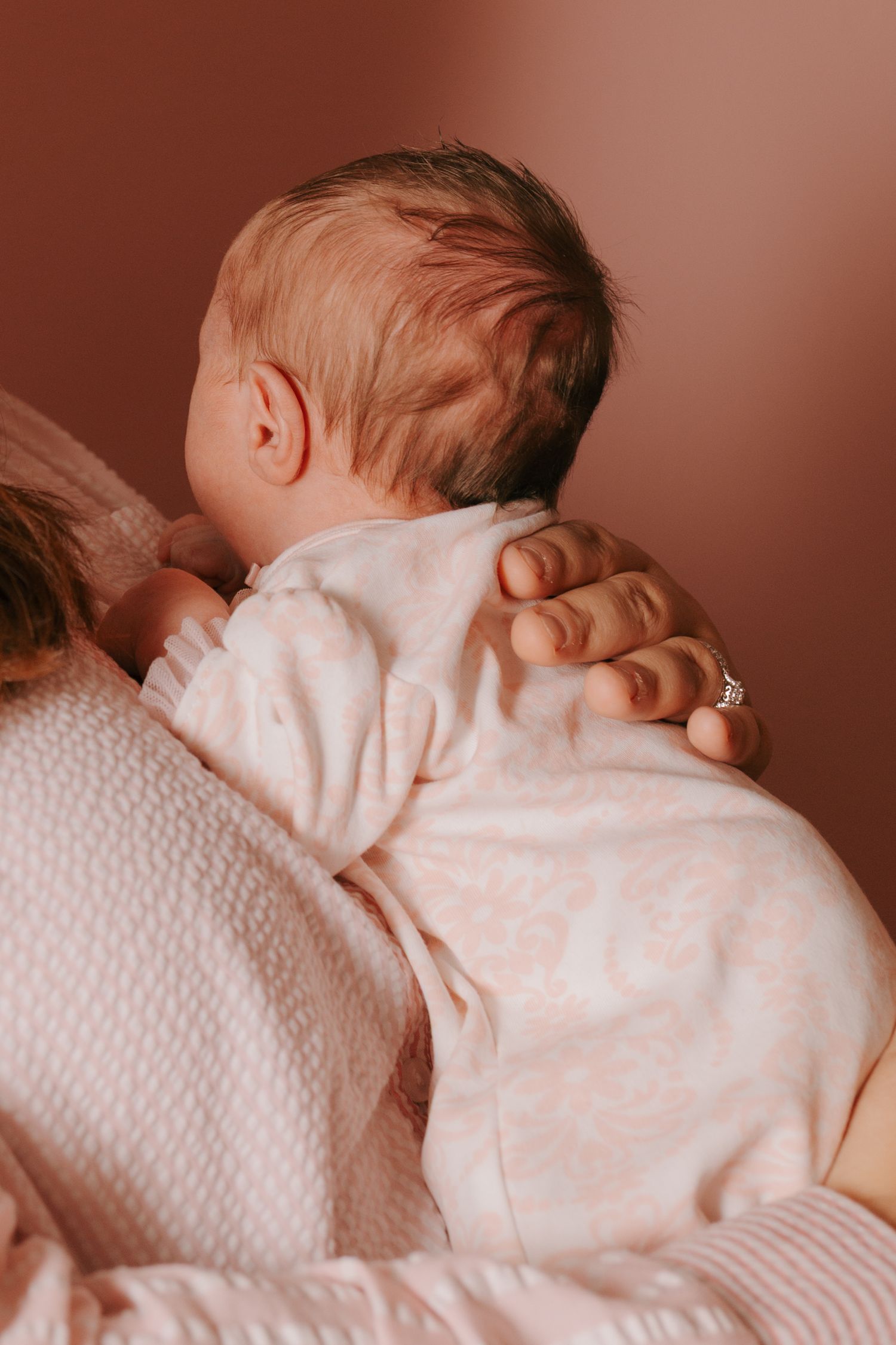 Mother gently holding newborn baby against her shoulder, wearing matching pink outfits in warm toned nursery.