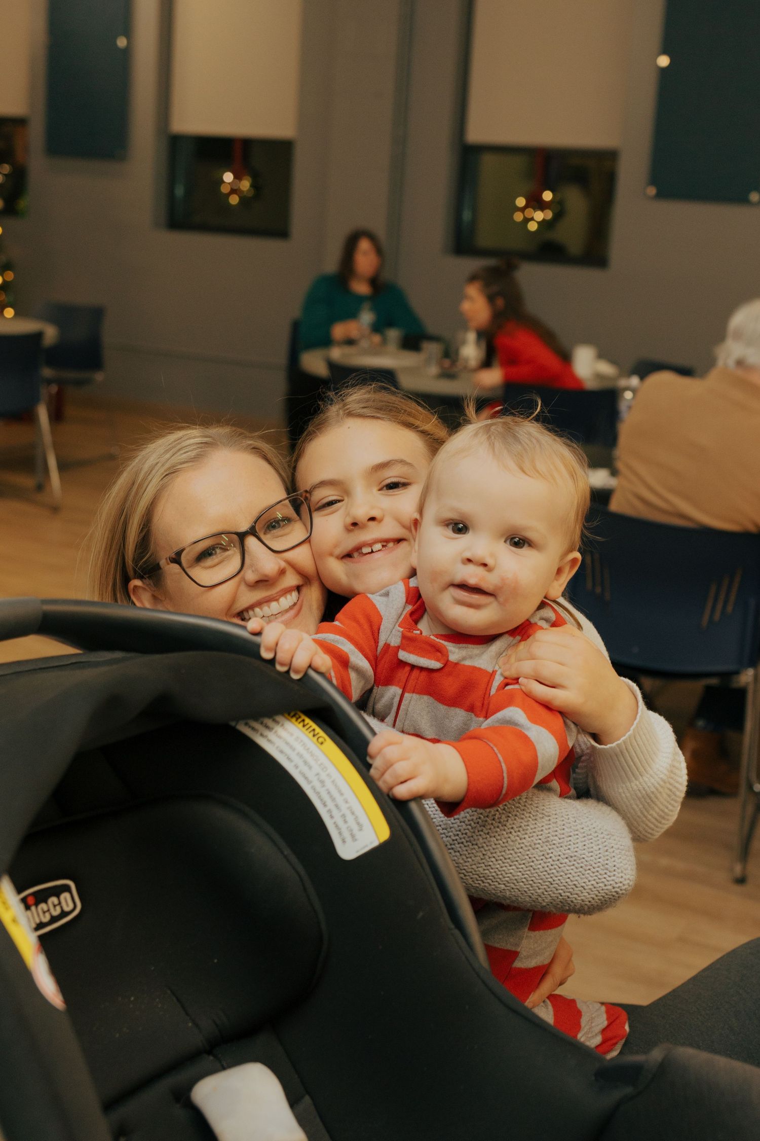 A woman with glasses smiles with two young children, including a baby in a striped outfit, at a holiday gathering.