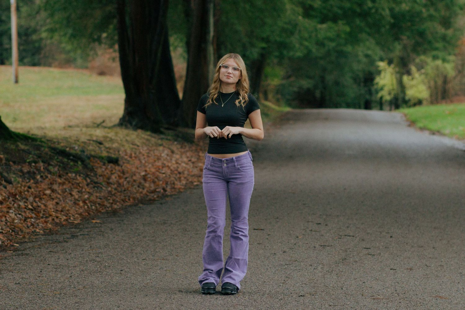 Young blonde woman in purple flare jeans standing on a tree-lined road in autumn.