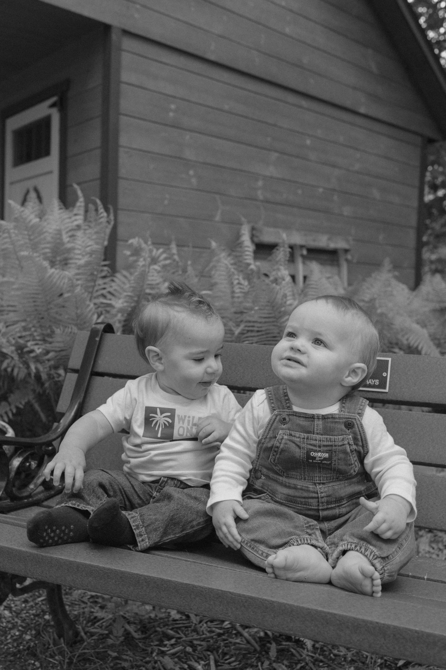 Black and white photo of two babies in overalls sitting together on a bench near a shed. 1st birthday session in Pittsburgh. Robin Hill Park