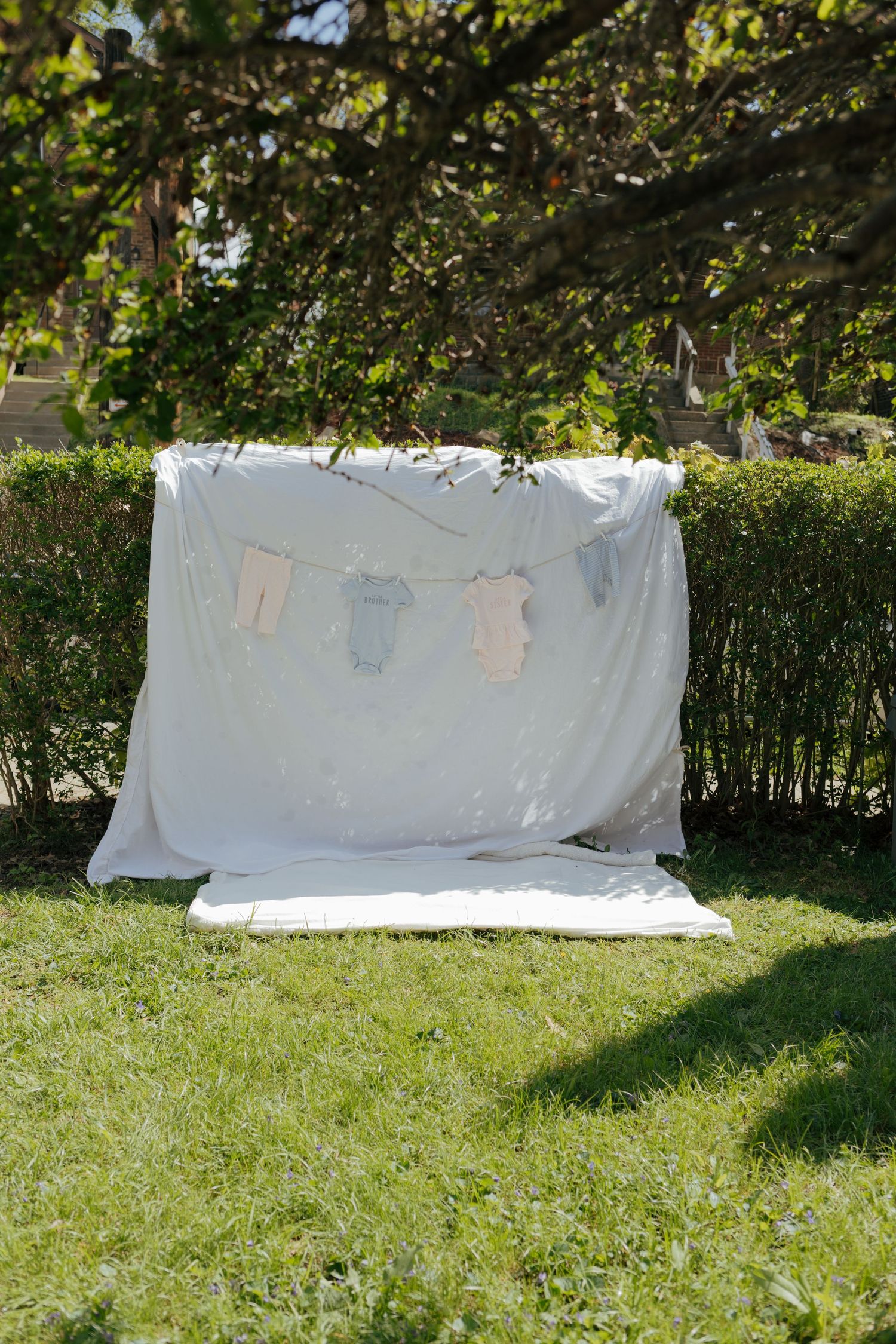 White fabric backdrop with mini baby onesies on clothesline in sunny outdoor garden.