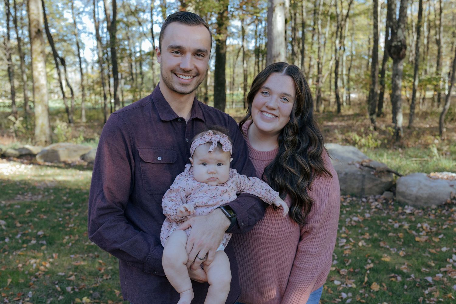 A smiling couple stands outdoors in a forest holding their infant daughter in pink clothing. Pittsburgh family session.