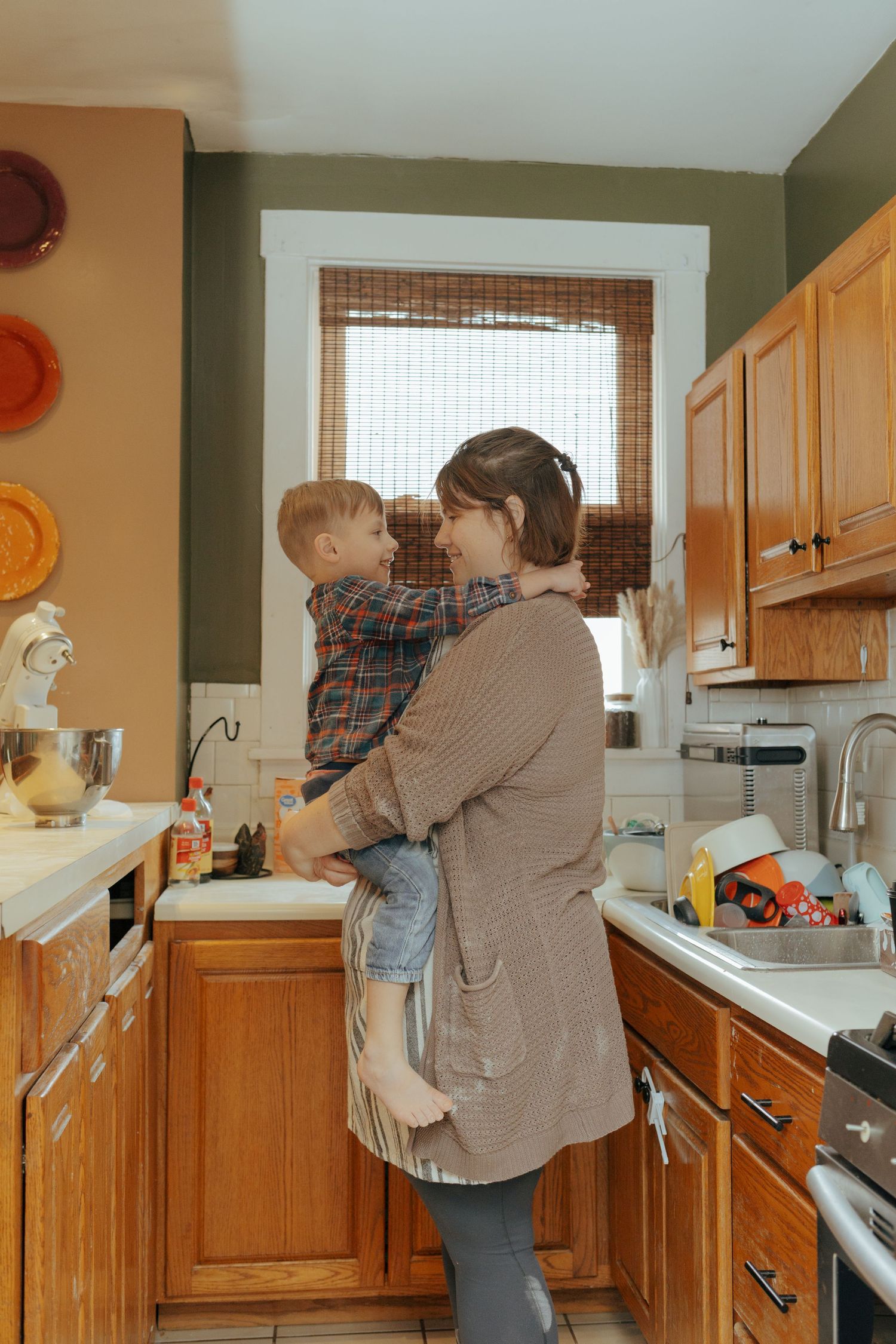 Mother holds toddler son in cozy kitchen with wooden cabinets and warm green walls in Pittsburgh