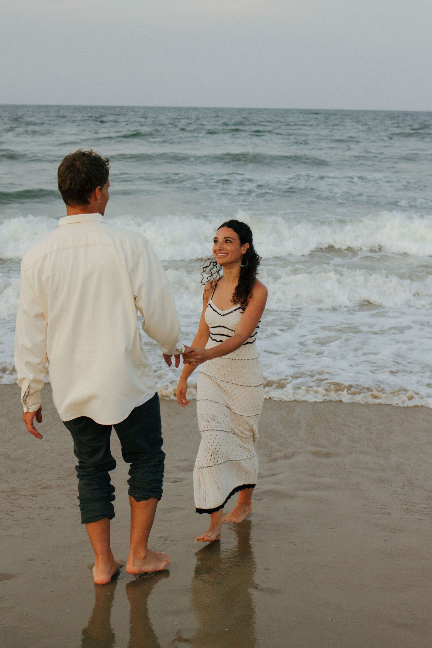 Couple holding hands on a sandy beach with ocean waves, woman smiling in a white knit dress.