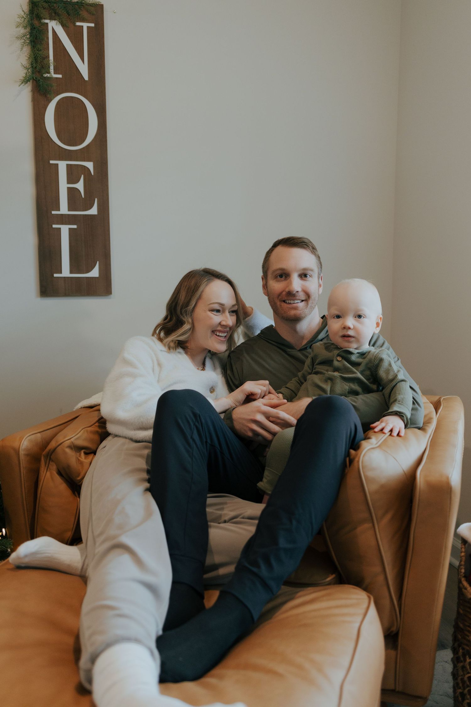 A happy family with a baby sits on a leather couch near a NOEL Christmas sign.
