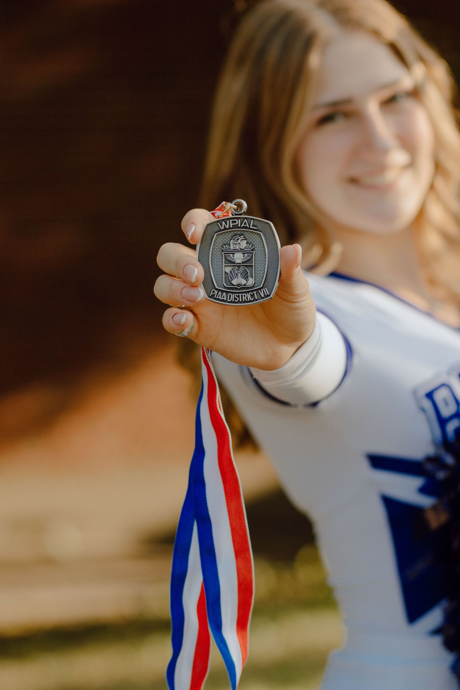 Smiling cheerleader holds up a silver championship medal with red, white, and blue ribbon toward camera.