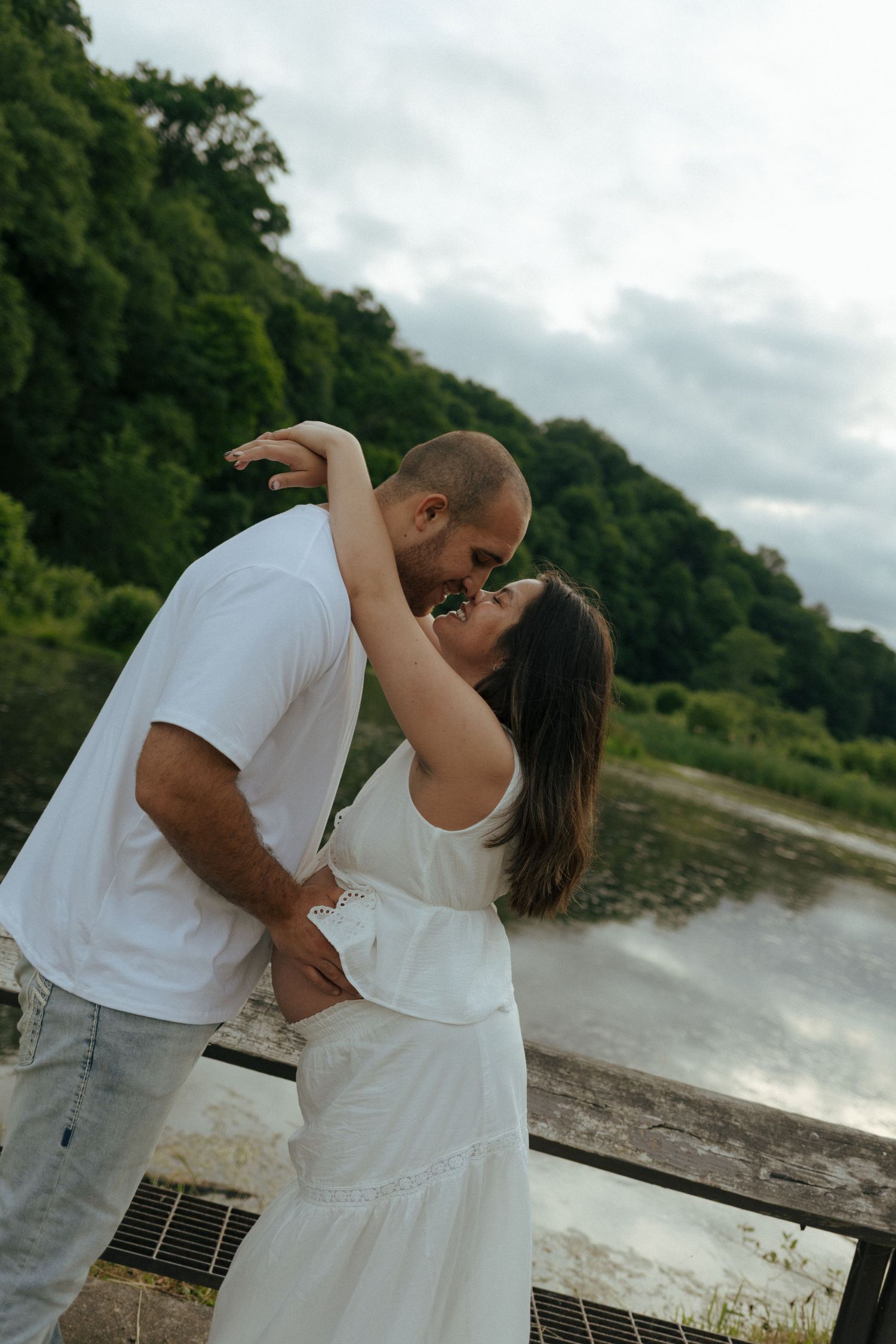 Couple in white outfits sharing a romantic moment near a scenic river with lush green trees in the background.