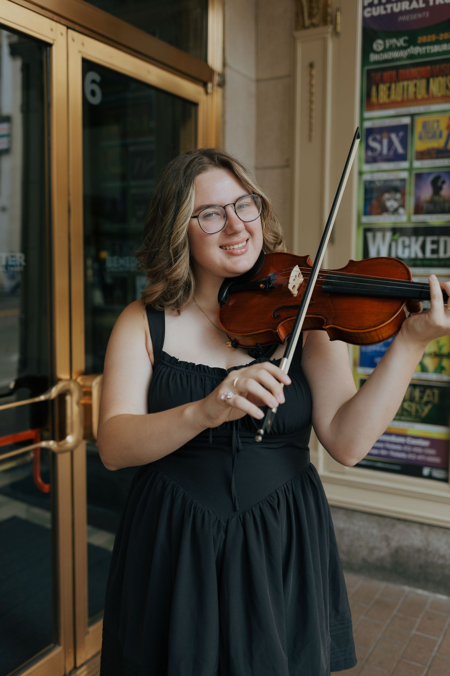 Smiling young woman plays violin outside theater displaying Broadway show posters including Wicked.