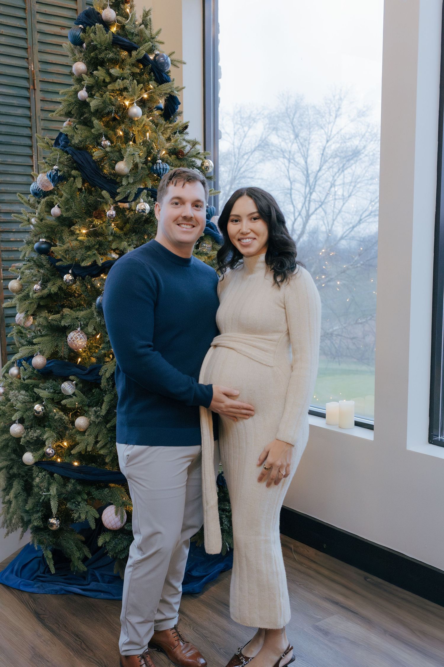 A smiling couple poses by a decorated Christmas tree, the woman wearing a cream dress showing her baby bump.