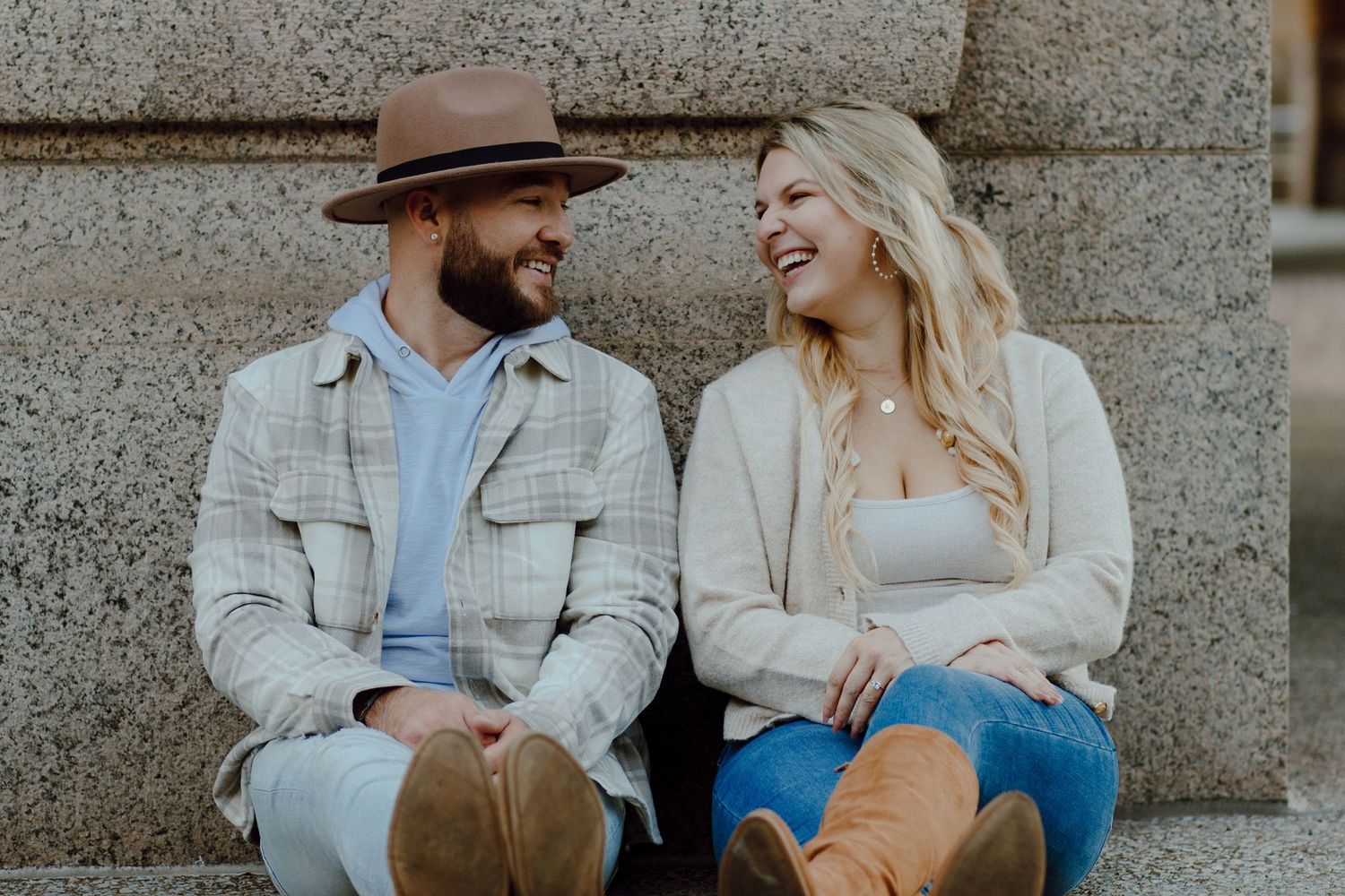 Happy engaged couple sitting on stone steps laughing together, man in plaid jacket and woman in cream sweater.