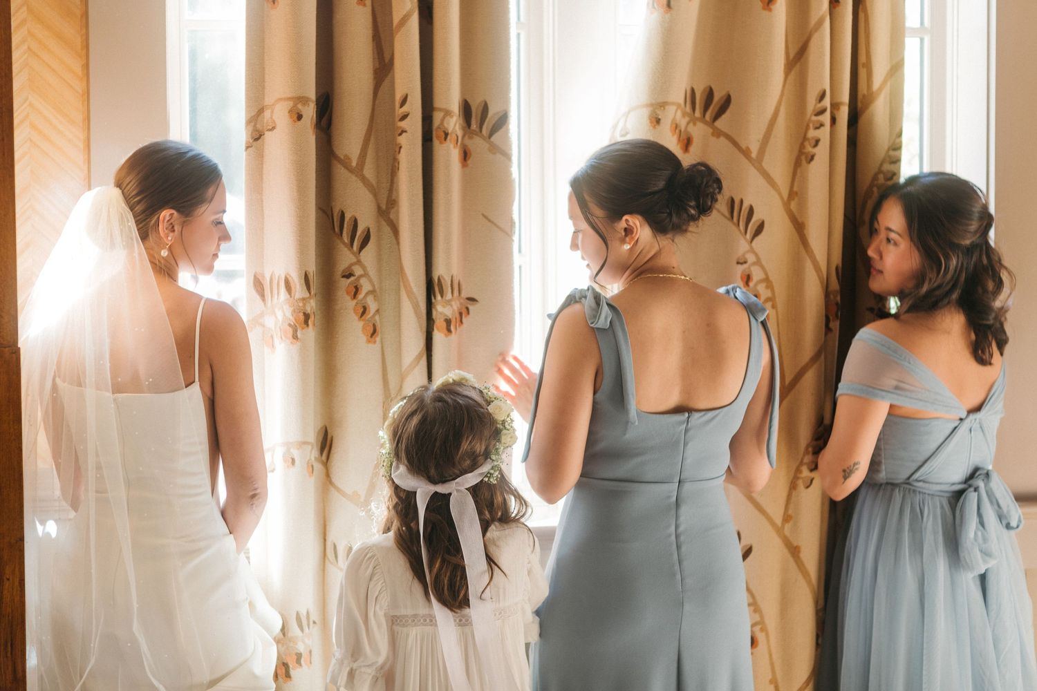 Bride in white gown and veil shares a tender moment with bridesmaids and flower girl by a sunlit window.