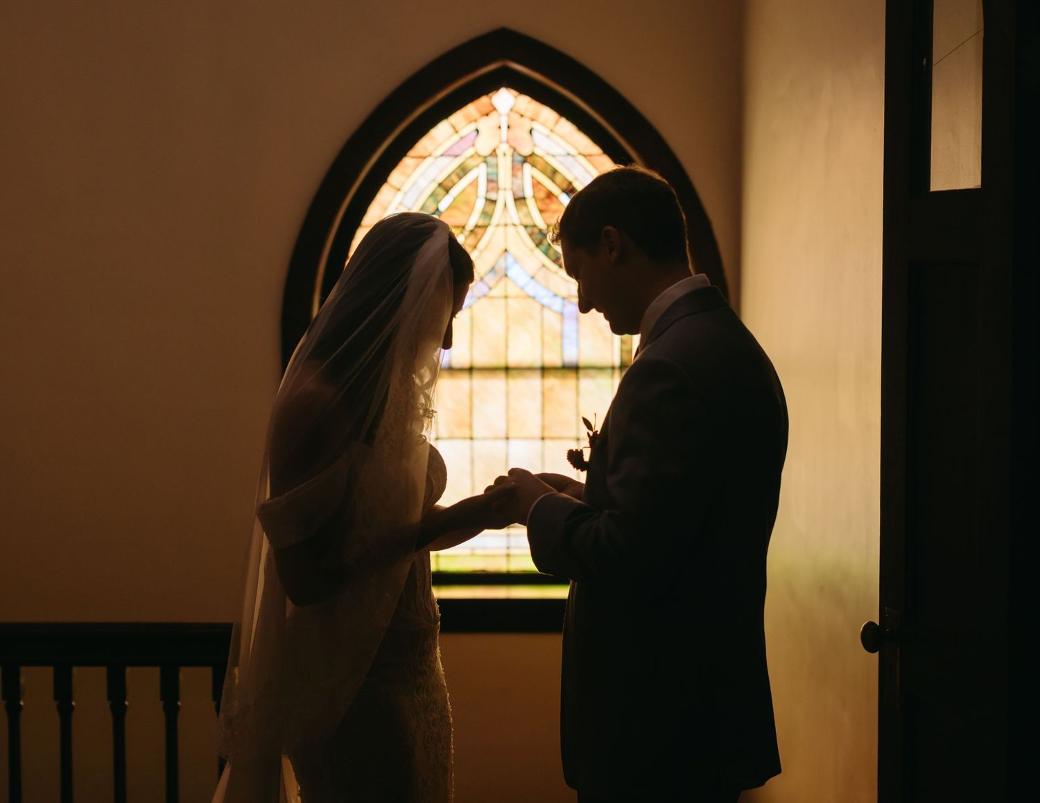 Bride and groom exchange rings in silhouette before a stained glass church window during their wedding ceremony.