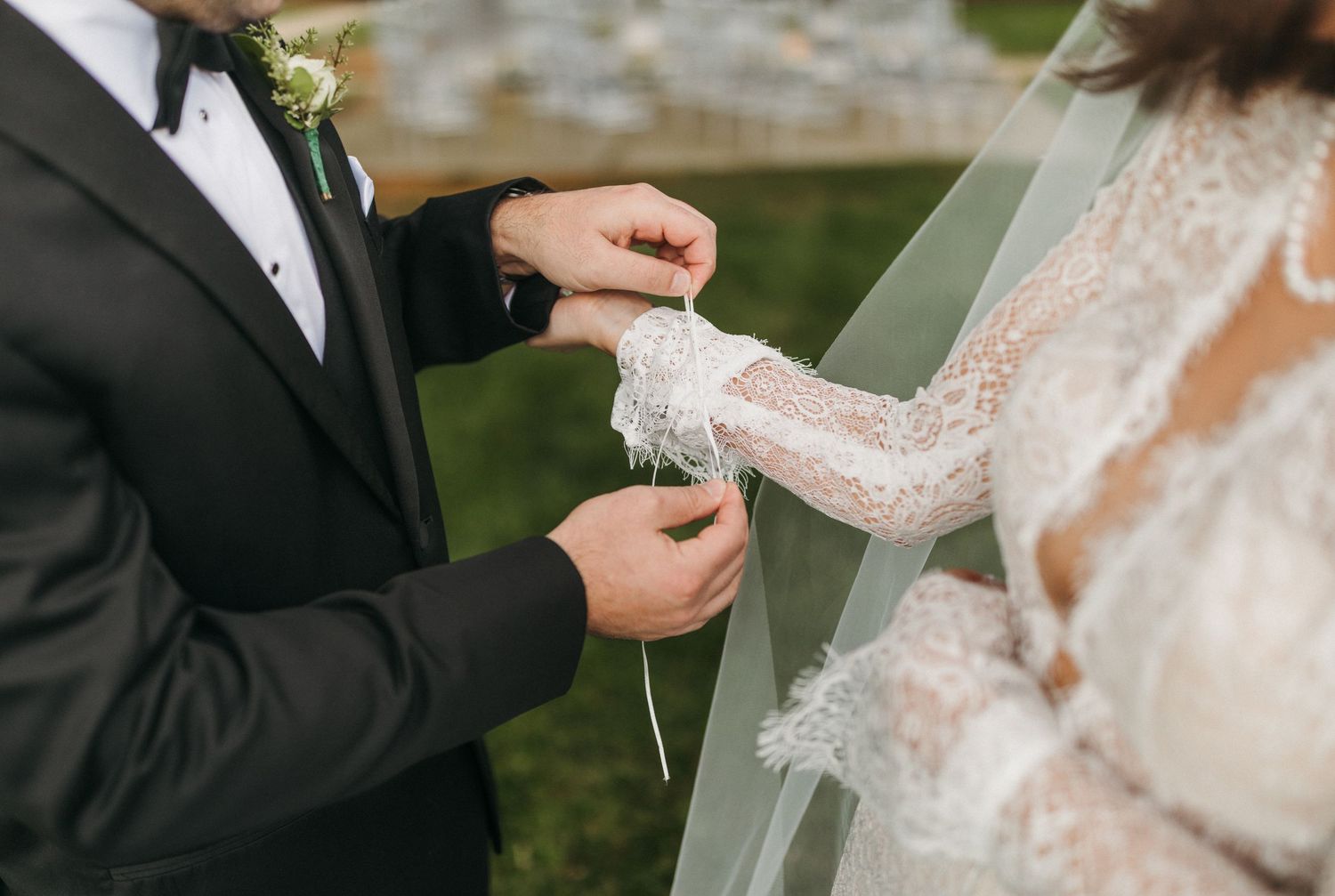 Groom adjusts lace cuff on bride's long-sleeve wedding dress during outdoor ceremony.