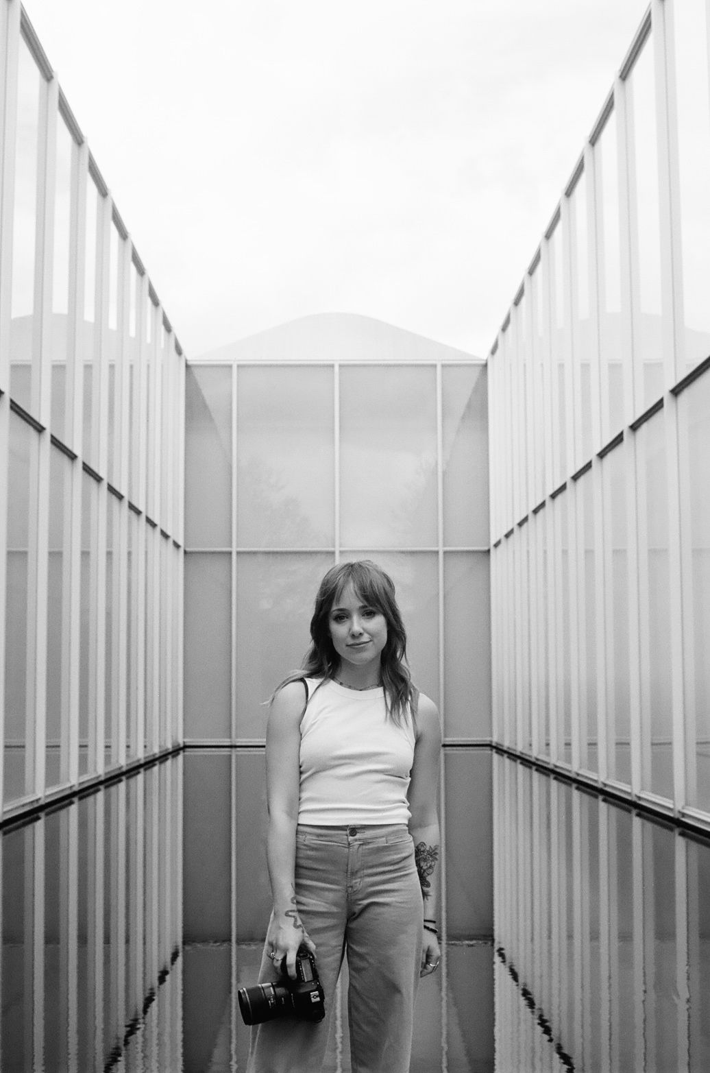 Black and white photo of a young woman holding a camera, standing in a symmetrical corridor with glass panels.