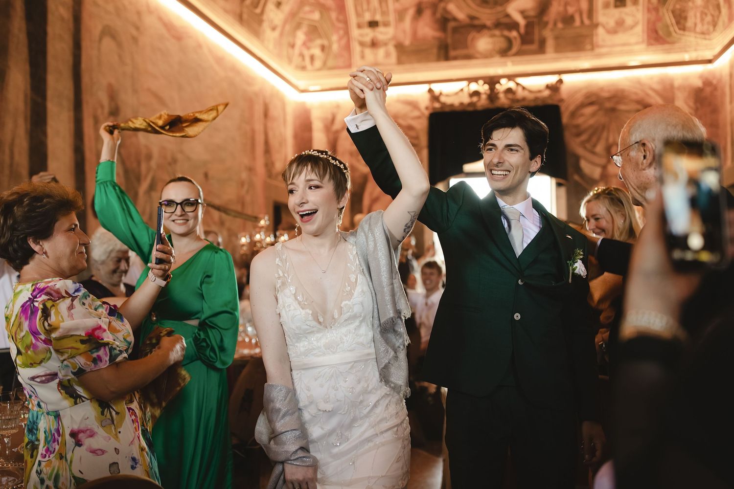 Excited bride and groom raising hands as they enter wedding reception surrounded by cheering guests.