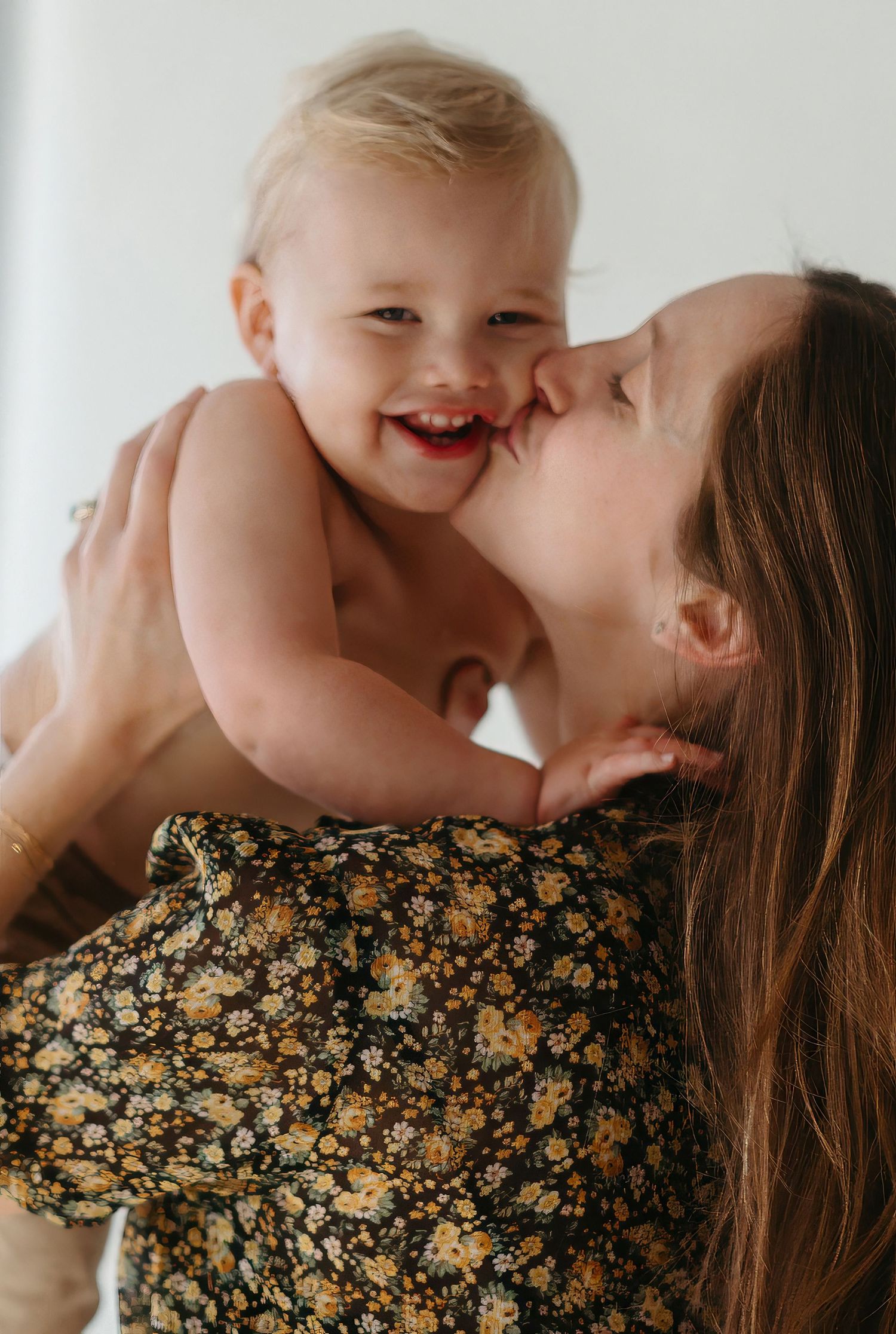 Happy laughing baby being kissed on cheek by mother wearing floral dress against white background.