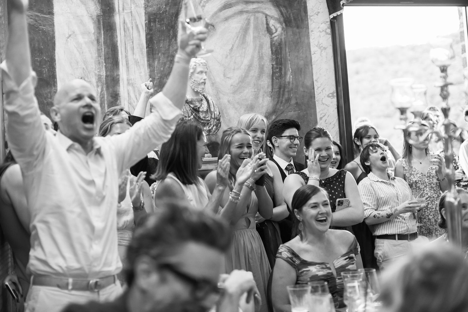 Black and white photo of joyful wedding guests celebrating and raising hands during reception in ornate venue.