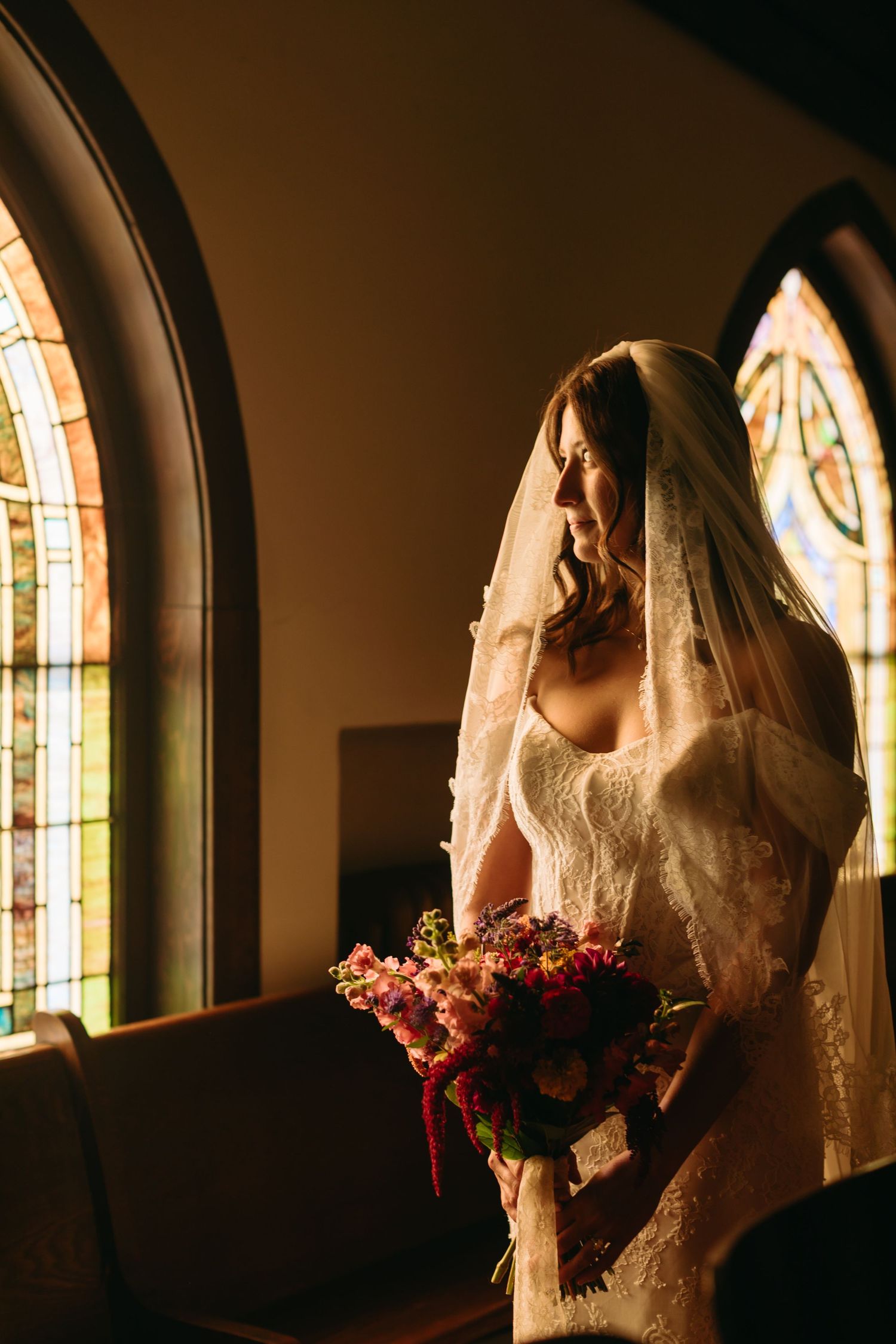 Bride in lace veil holding colorful bouquet by stained glass church windows in warm light.