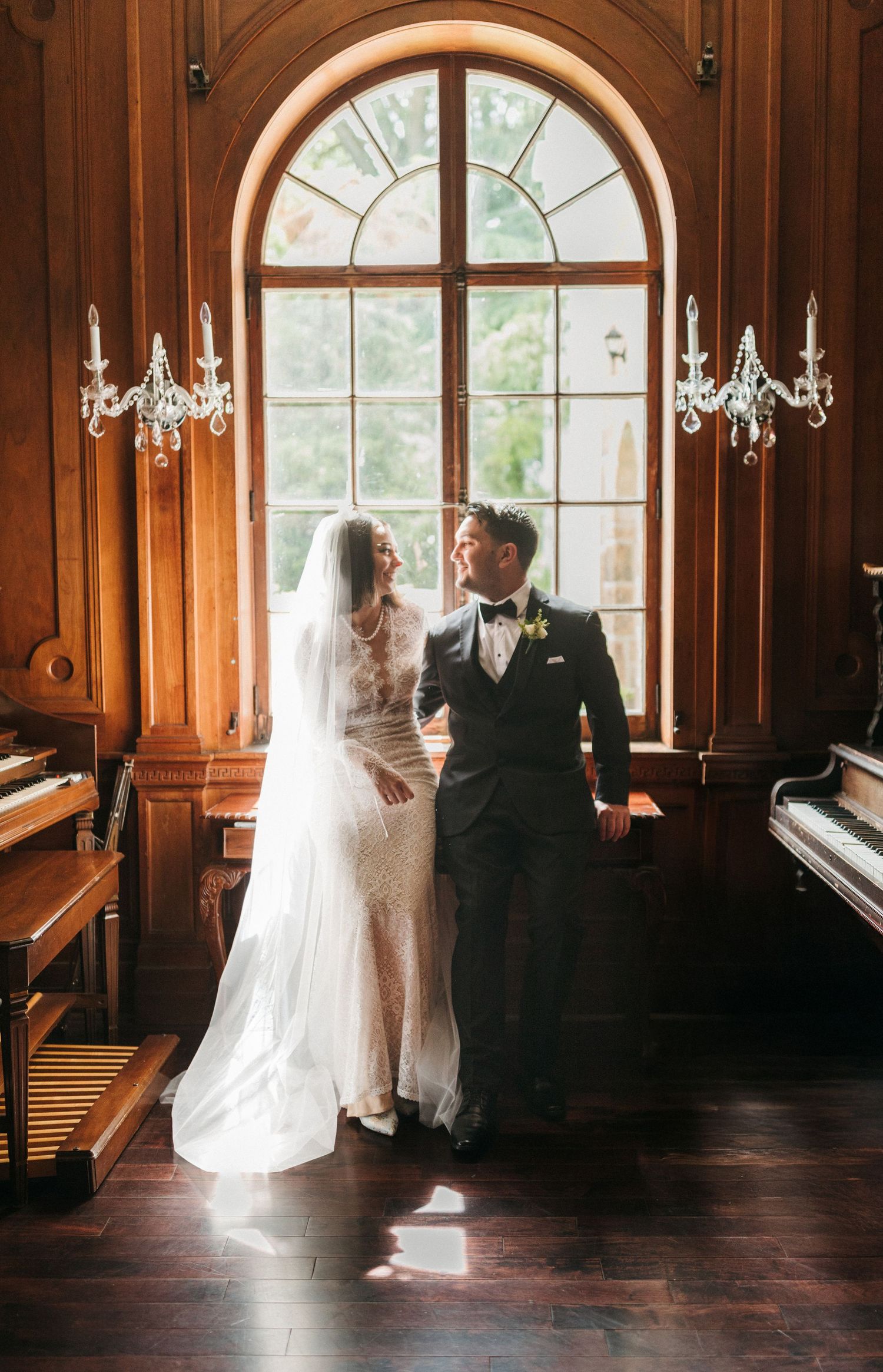 Bride and groom share romantic moment by arched window in wood-paneled room with chandeliers.