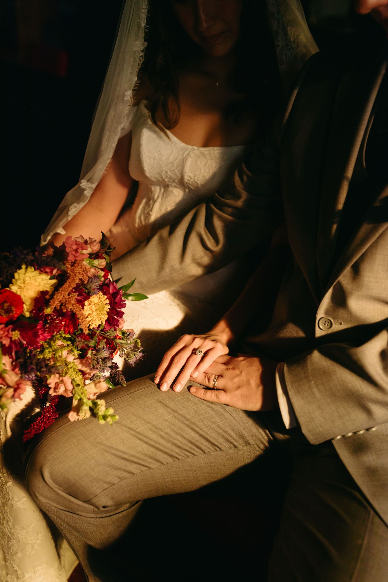 Bride and groom holding hands with wedding rings visible, seated together with a colorful bridal bouquet in warm golden light.
