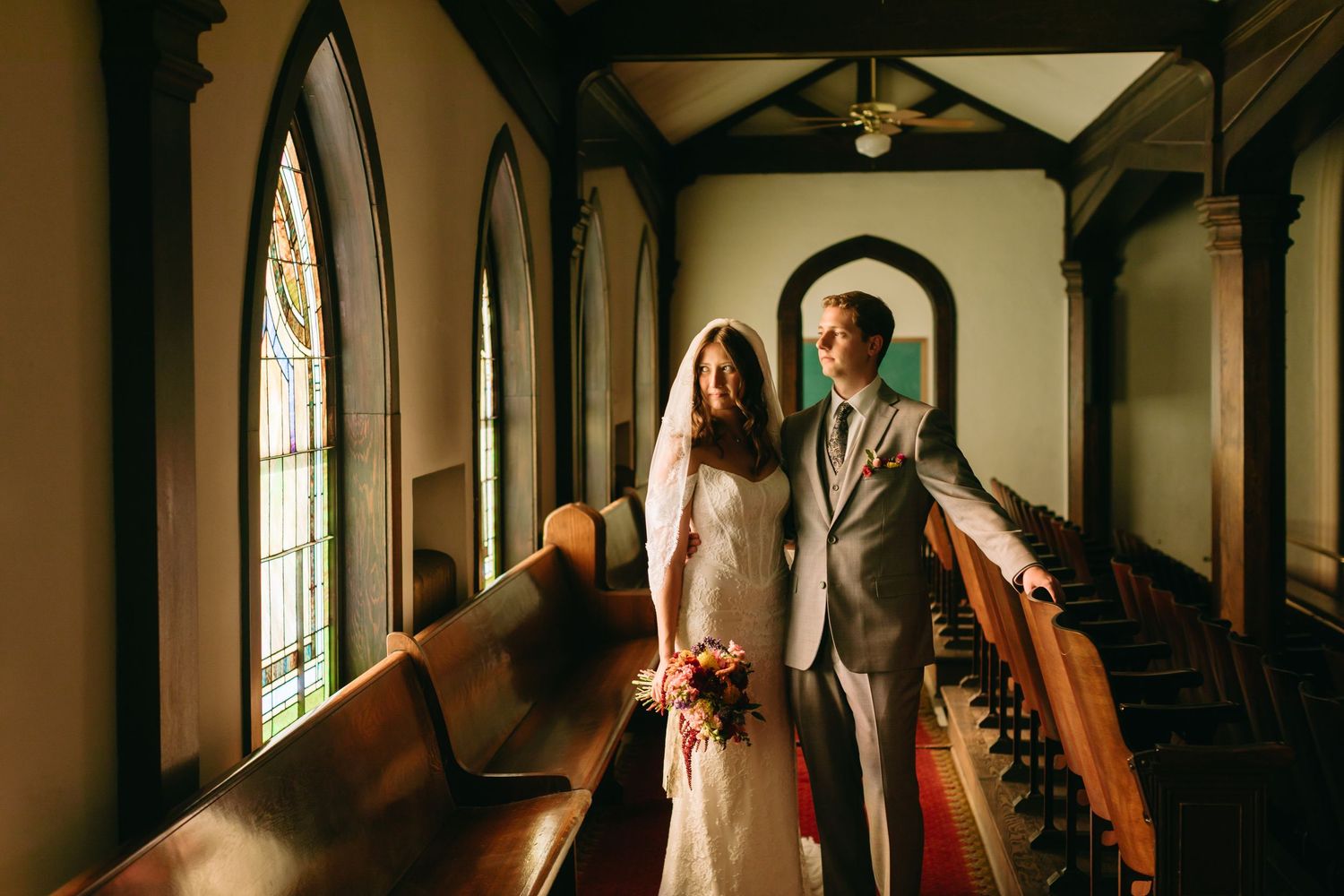 Bride and groom stand together in sunlit chapel between wooden pews.