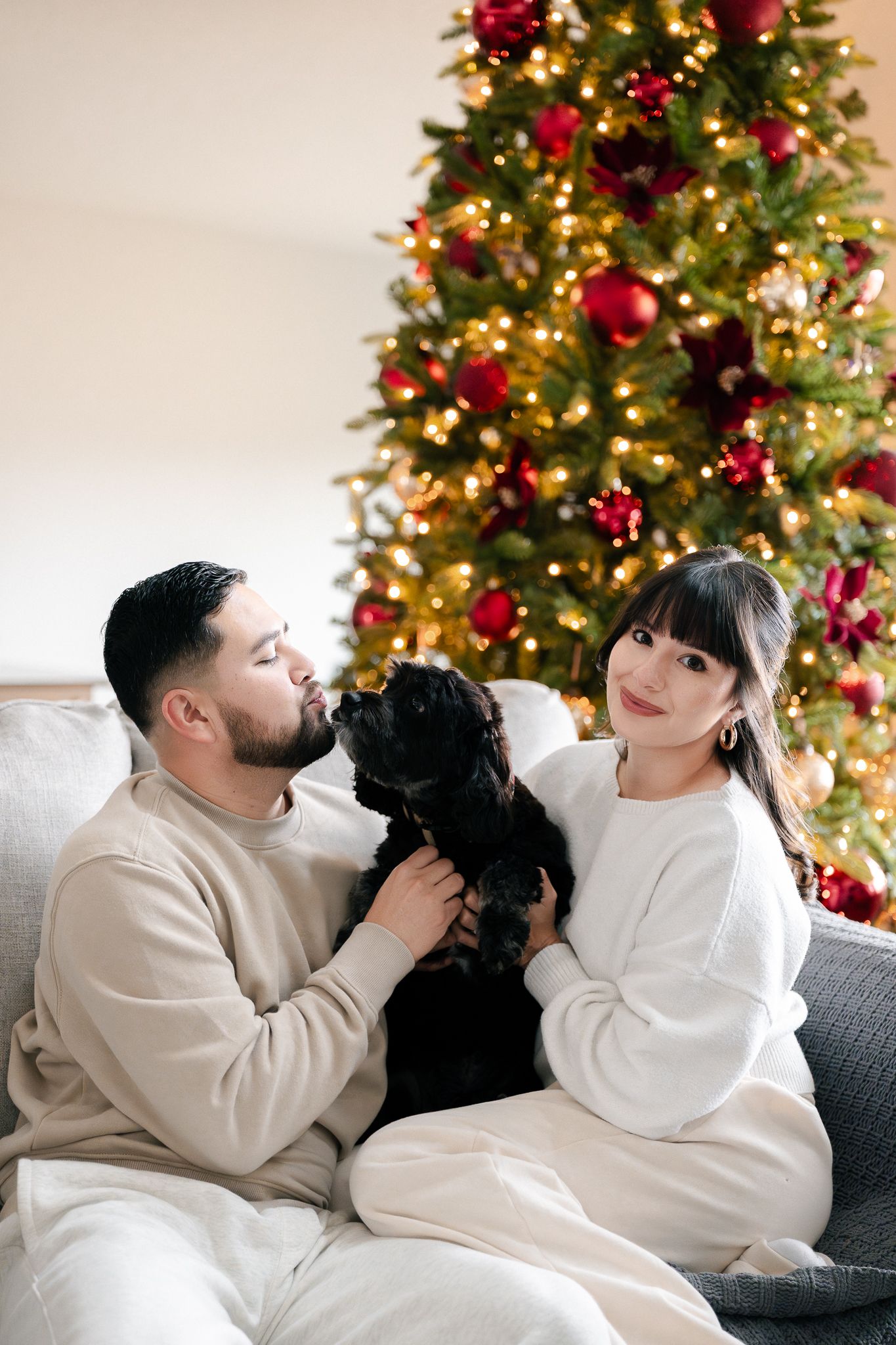 A couple in matching white sweaters cuddle with their black dog on a couch in front of a decorated Christmas tree.