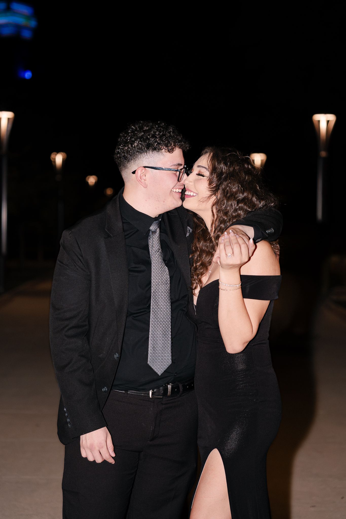 A couple dressed in formal black attire share an intimate moment at night under outdoor lighting.